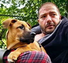 A man lying outdoors with a brown dog resting on his lap, surrounded by green trees, during daytime.