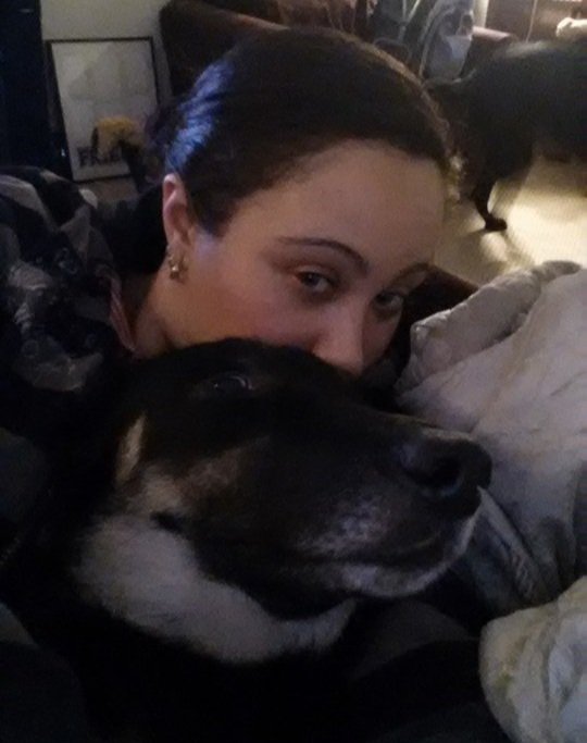 A woman with dark hair and earrings cuddling with a large, black and tan dog on a couch inside a cozy room.
