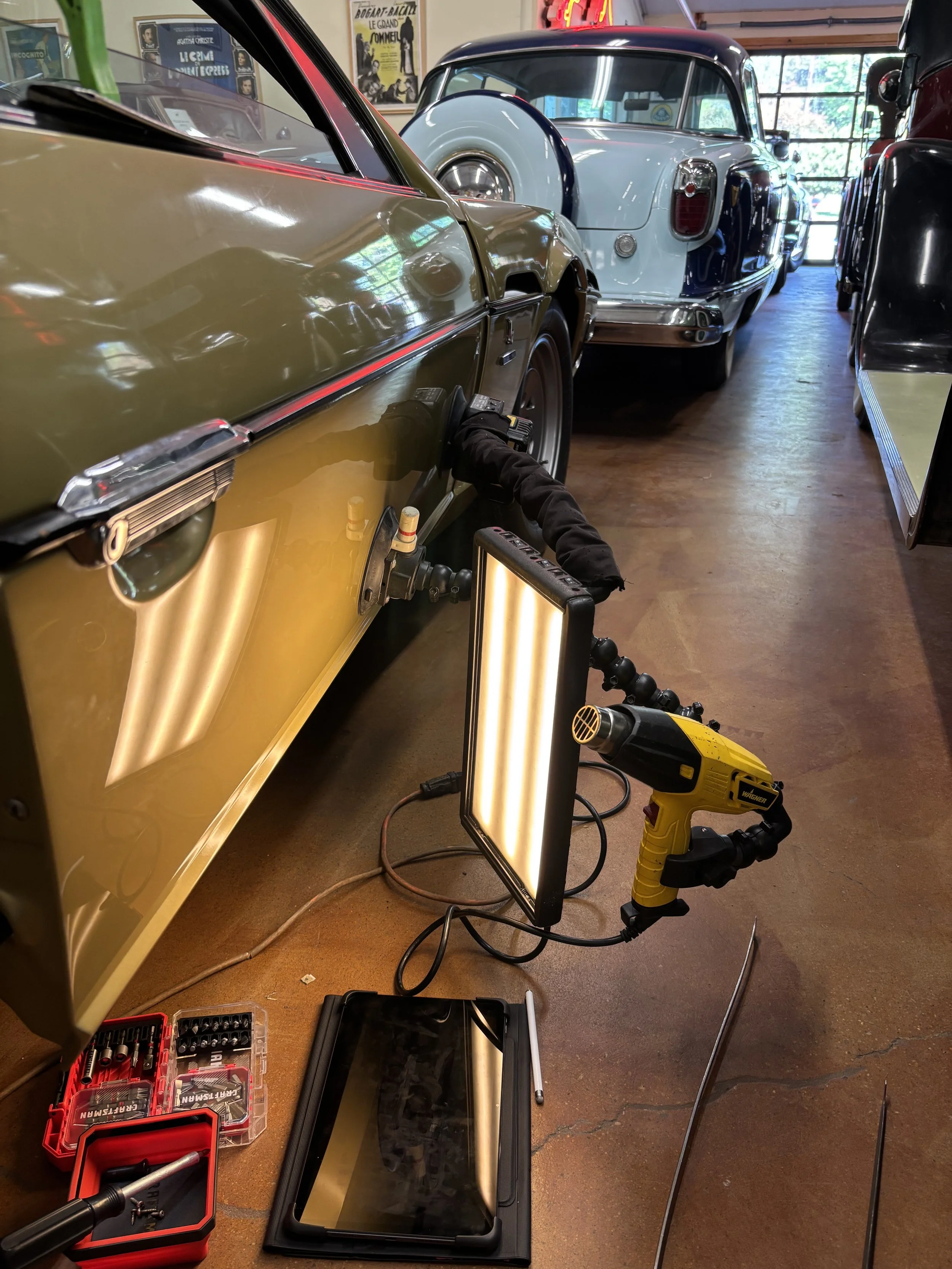 A green classic car undergoing repair with tools and equipment nearby in a garage setting. A light, heat gun, and various tools are seen beside the car. Other vintage cars are visible in the background.