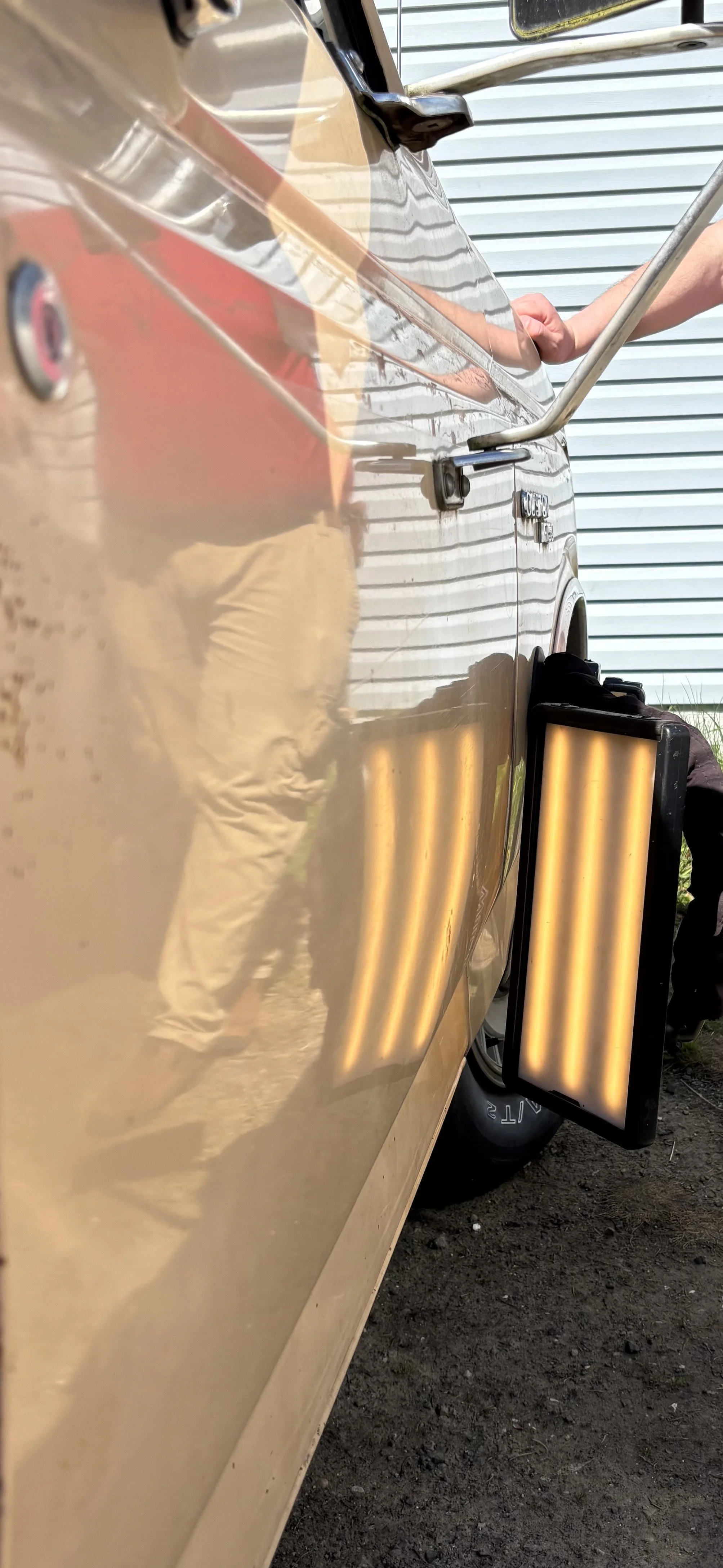 Close-up of a classic car door with a reflection of a person wearing khaki pants. A paintless dent repair light is used to highlight dents on the car surface. Vehicle background includes a garage door.