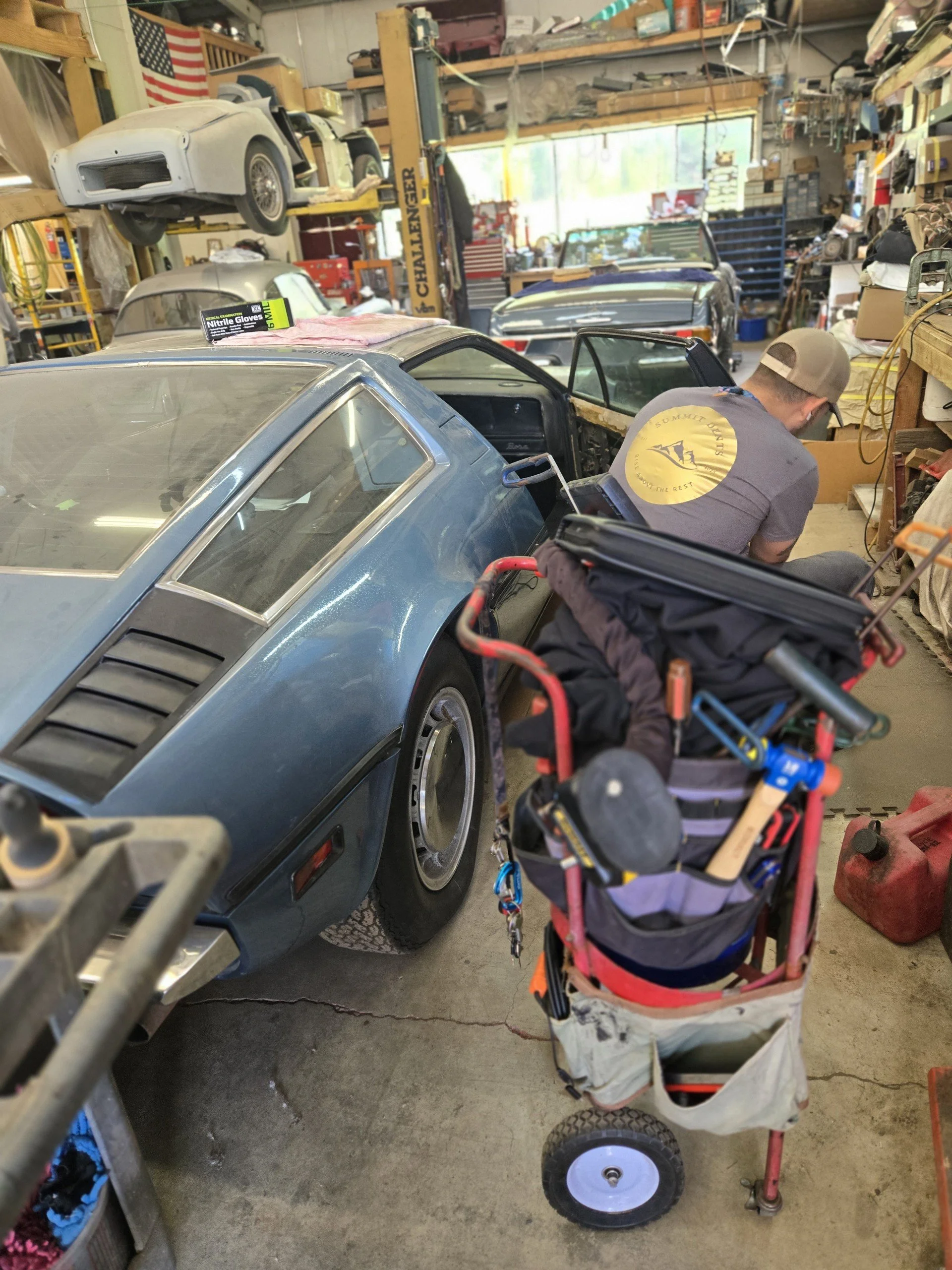 Man working in auto repair shop, surrounded by vintage cars and tools.