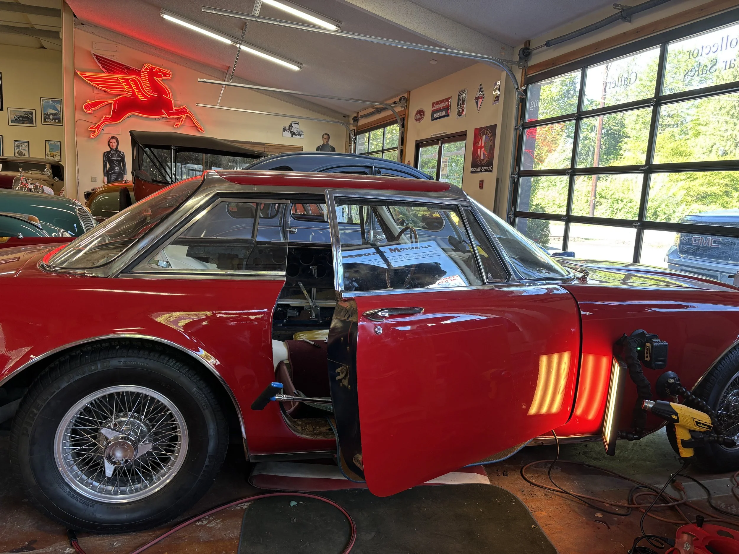 Vintage red car in a garage with open doors, showcasing wire wheels. Neon Pegasus sign and classic car memorabilia decorate the walls. A GMC vehicle is visible outside through the glass doors.