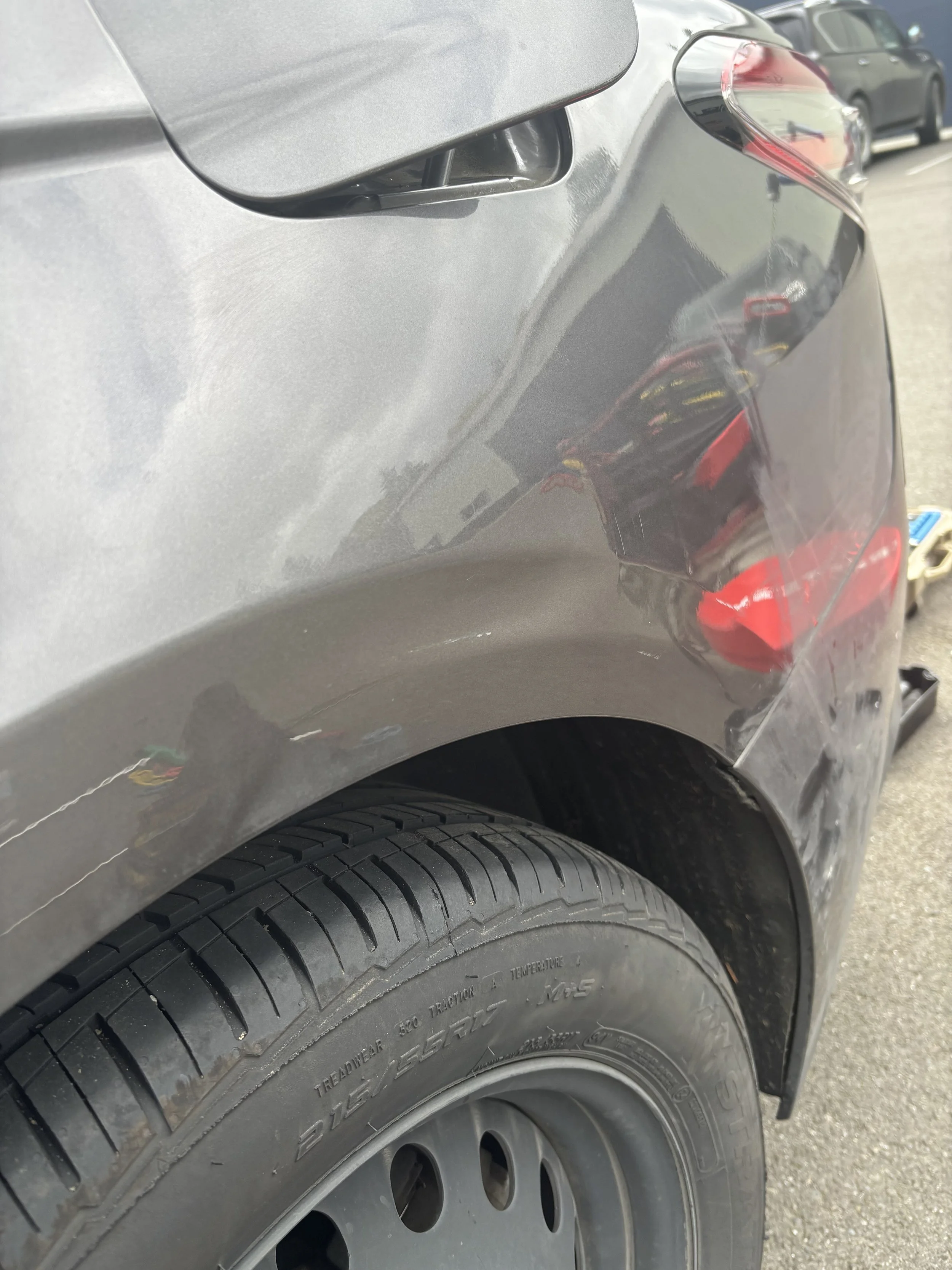 Close-up of a dark-colored car showing a rear tire, wheel arch, and part of the rear bumper. There are visible scratches and damage on the bumper.
