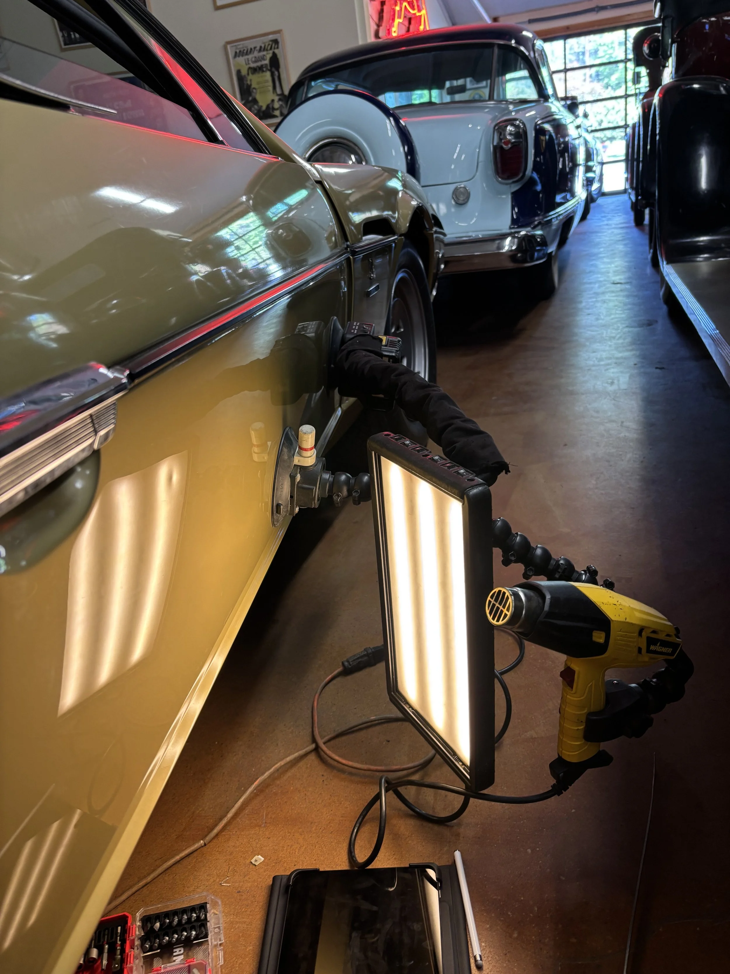 Workshop with classic cars, showing a close-up of a green car's panel under repair with a light fixture and heat gun attached. Other classic cars are visible in the background.
