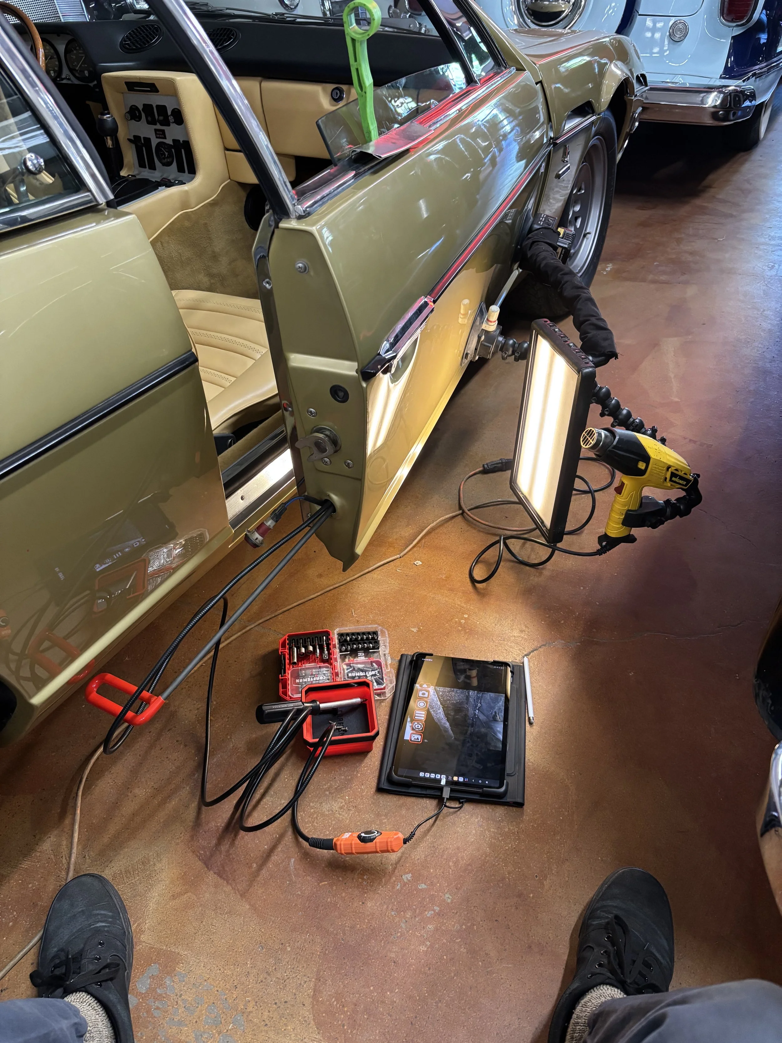 Car repair setup showing an open door, inspection lights, toolkit, and tablet on the floor of a garage.