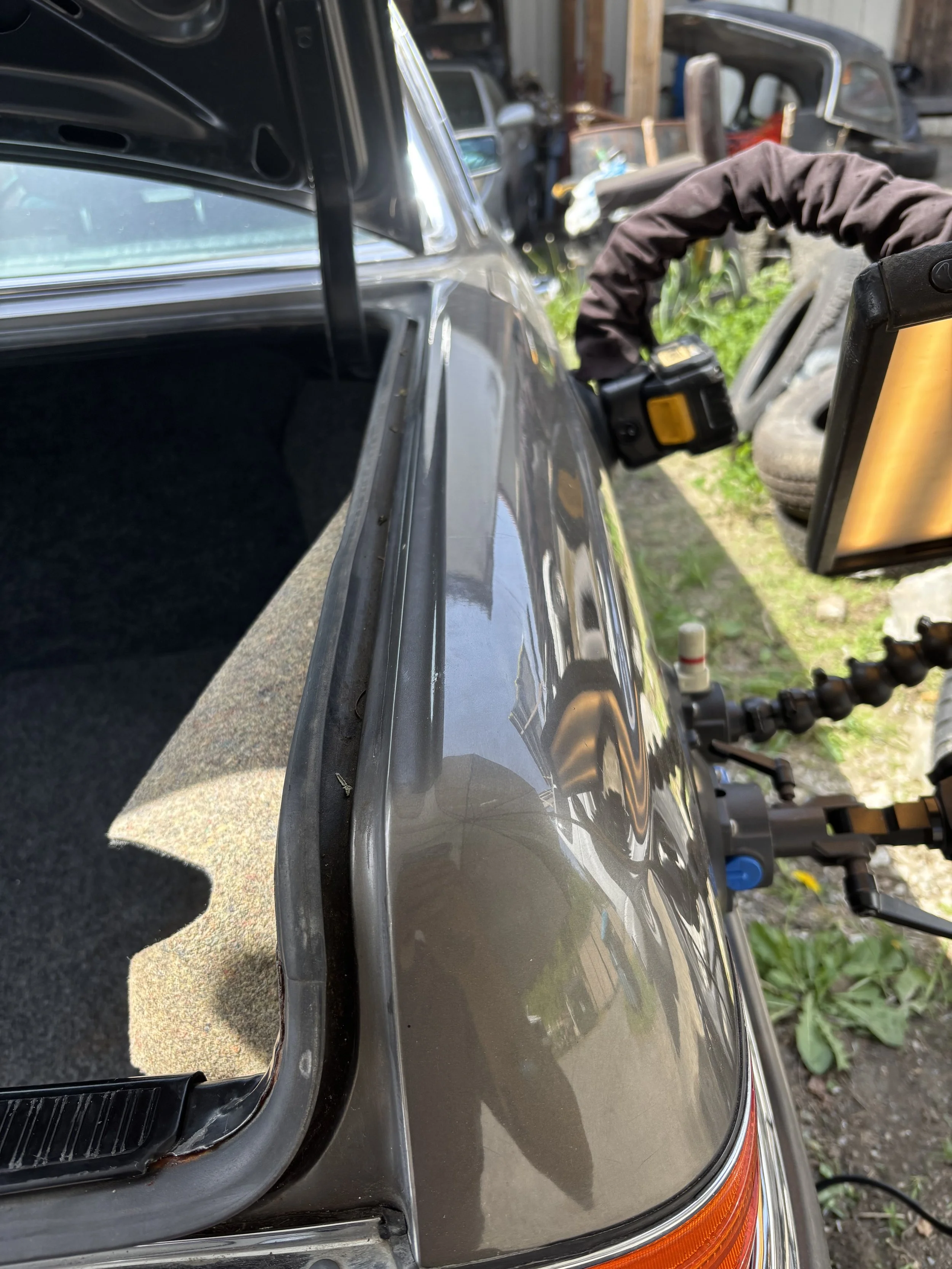 Close-up of a car's open trunk and rear panel with a reflective surface, showing part of the tail light. There is equipment attached nearby, possibly for repair or inspection.