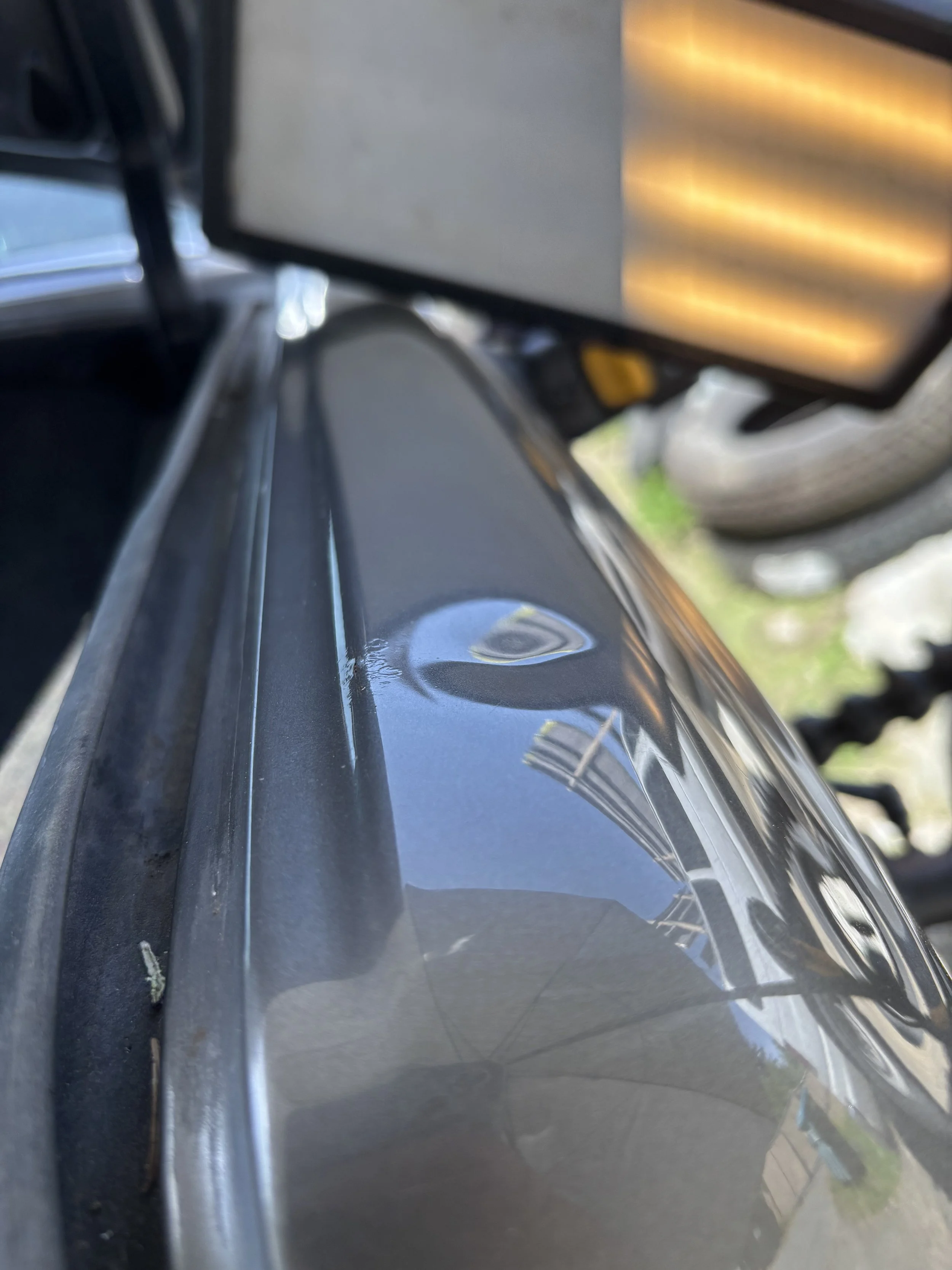 Close-up of a car door with a visible dent, reflecting a nearby building and umbrella, with a blurred background and a light source.
