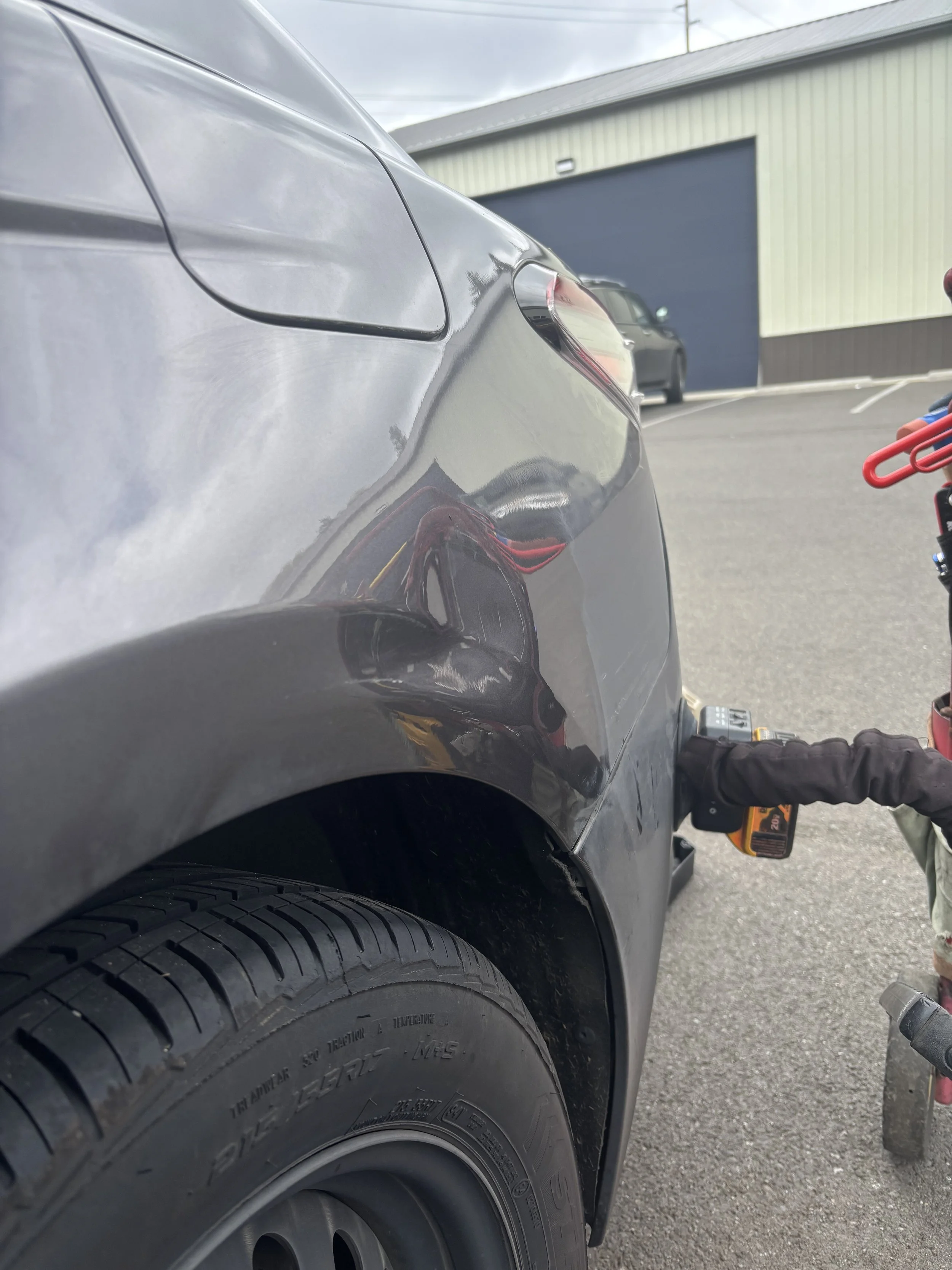 Close-up of a car's rear quarter panel and tire with a visible dent. A tool is positioned near the dent, likely for repair. A building and another car are visible in the background.