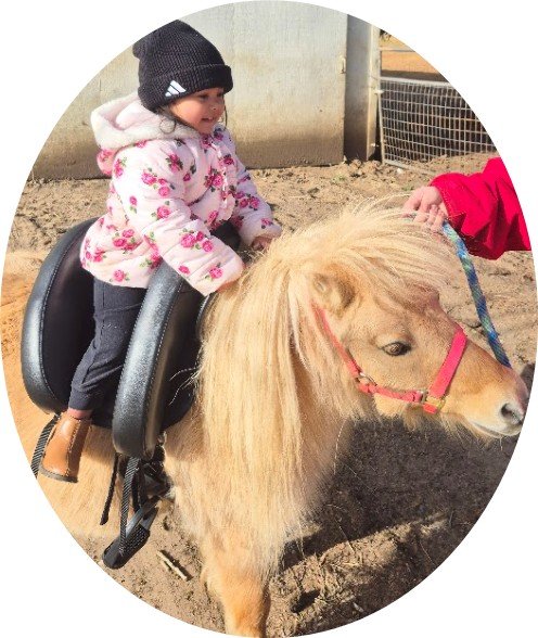 A young girl wearing a black beanie and floral jacket rides a small light-colored pony with a red bridle, while someone holds its lead rope in a horse pen.