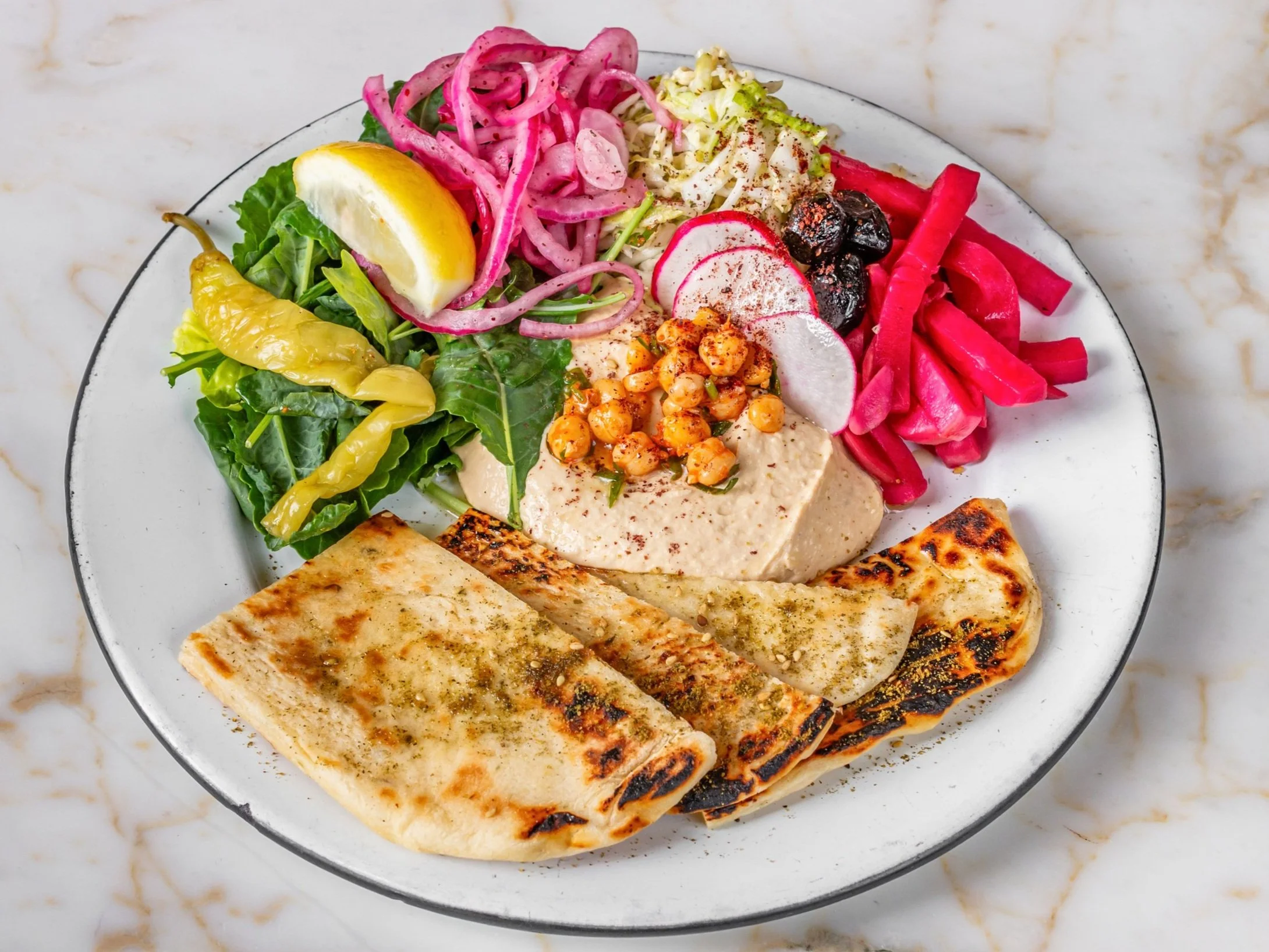 A white plate with Mediterranean food including grilled flatbread, chickpea hummus topped with paprika, pickled vegetables, olives, radish slices, cucumber, greens, and lemon wedge.