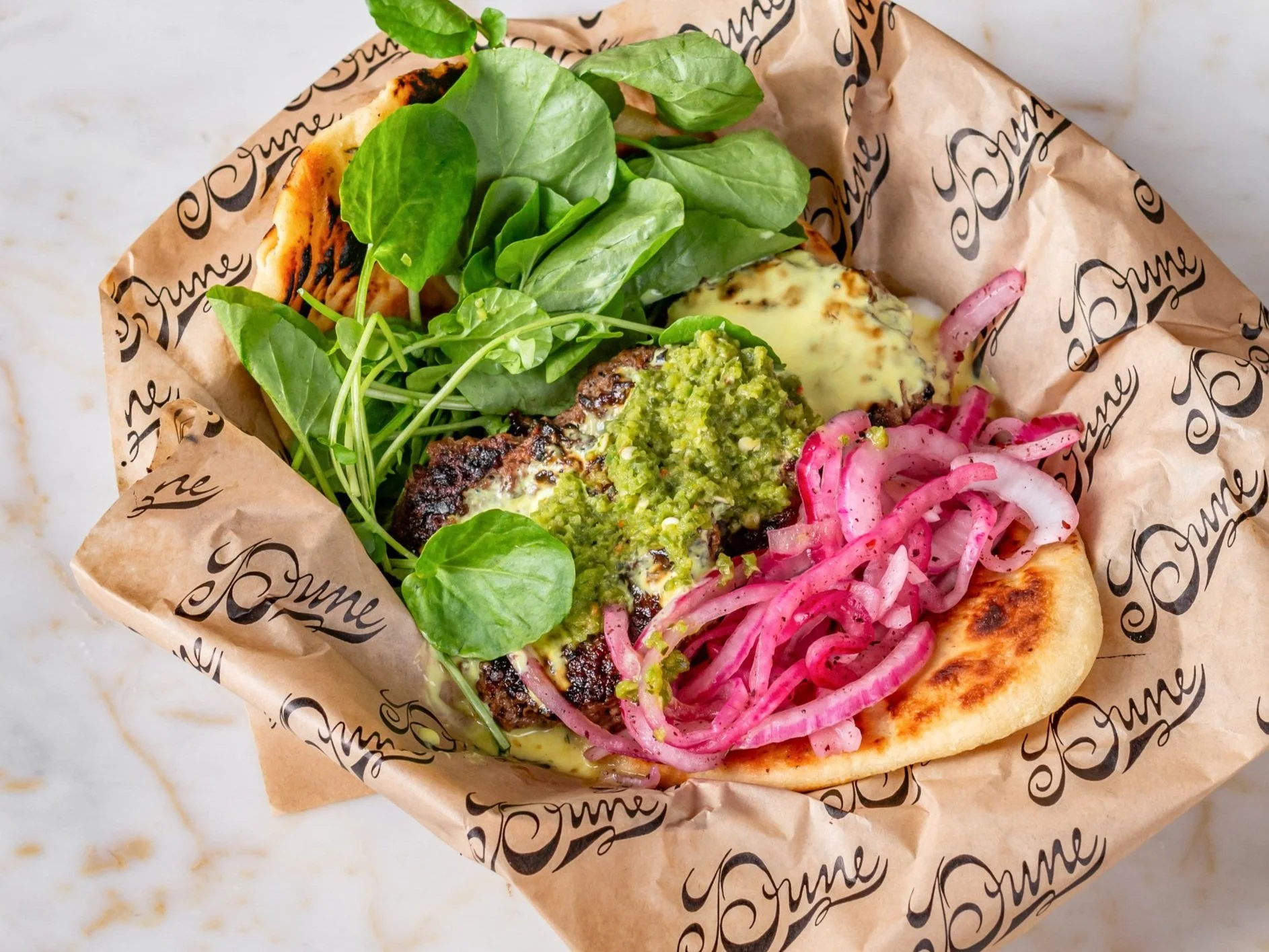 A gyro sandwich wrapped in branded parchment paper containing lettuce, grilled meat, green sauce, pickled onions, and spinach leaves.