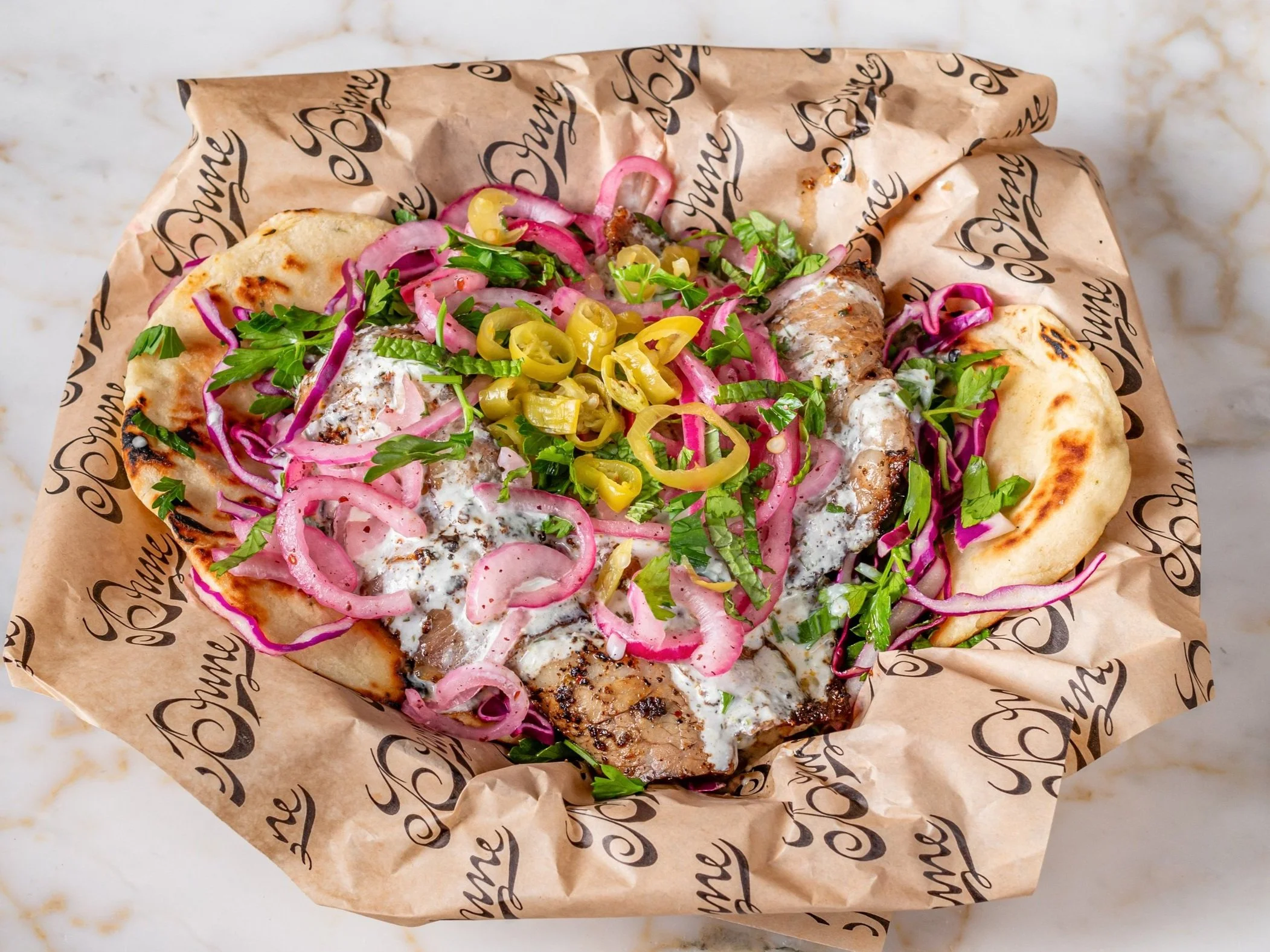 A pita bread filled with grilled chicken, topped with chopped herbs, pink pickled onions, and sliced yellow chili peppers, served on branded parchment paper.