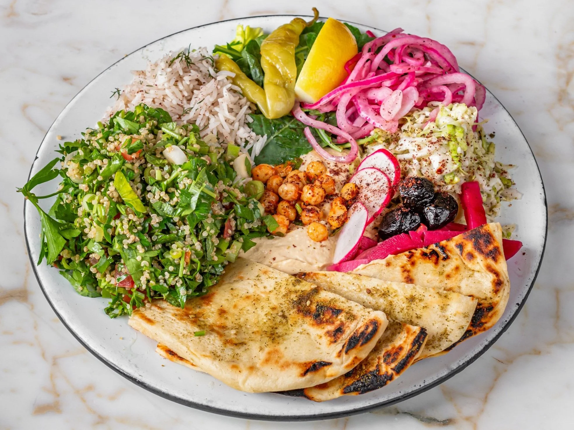 A plate with rice, kale salad, grilled flatbread, chickpeas, sliced radishes, pickled onions, hummus, black olives, and pickled vegetables on a marble surface.