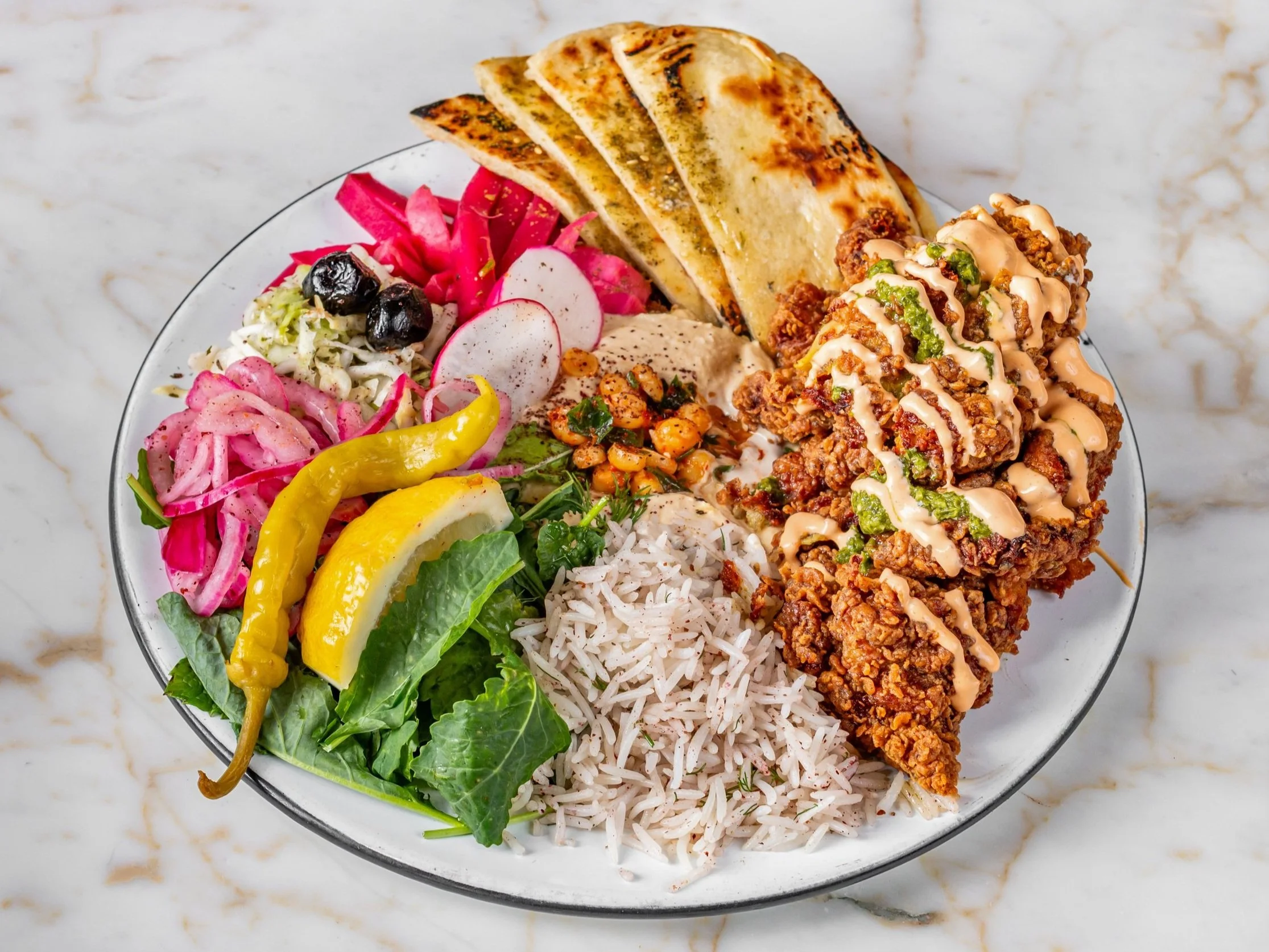 A plate of Mexican food including fried chicken with sauce, rice, grilled flatbread, a salad with pickled vegetables, lemon wedge, peppers, radishes, olives, and black beans.