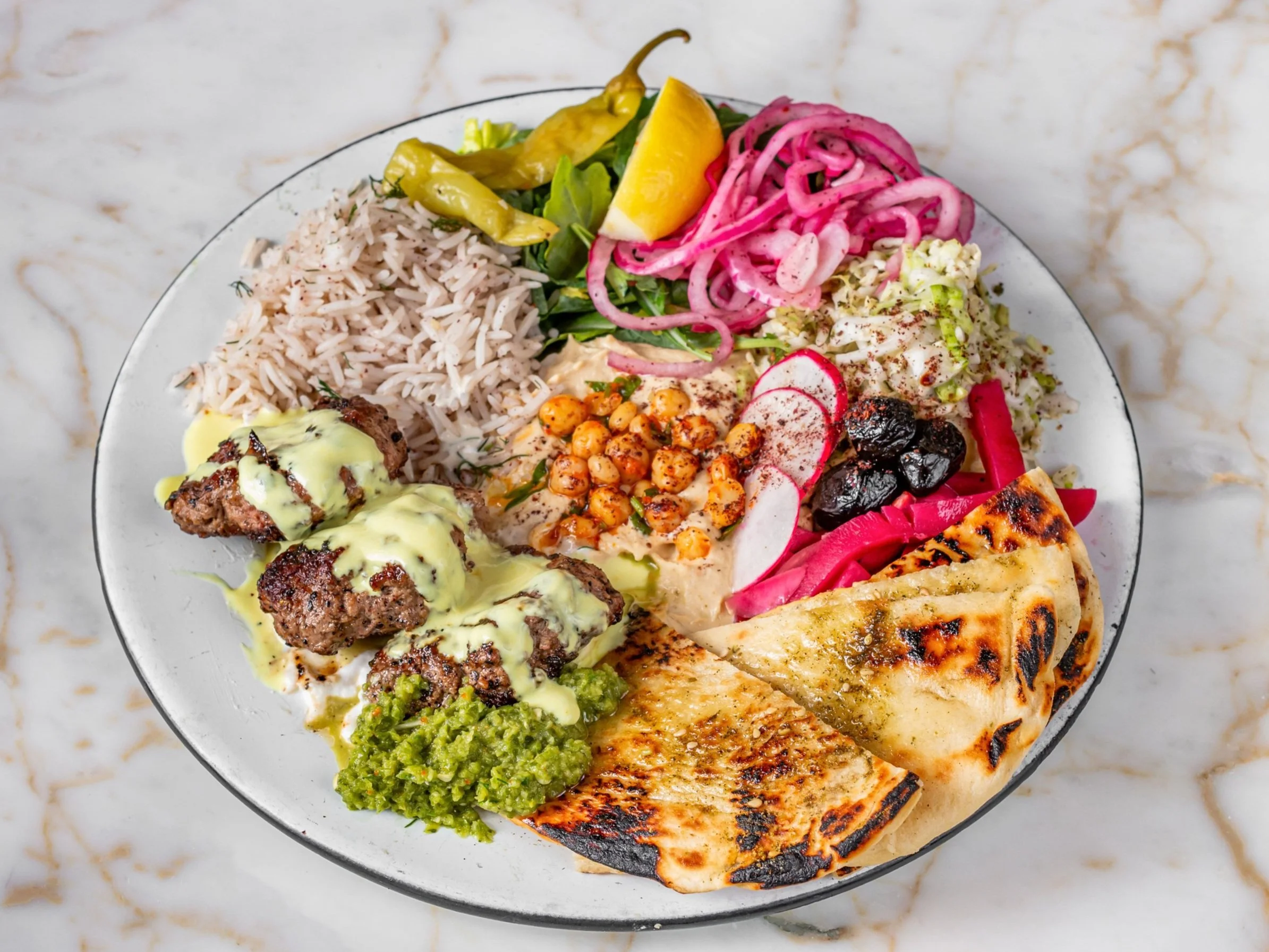 A plate of Middle Eastern cuisine featuring rice, grilled meat with sauce, hummus, flatbread, tabbouleh, pickles, fresh vegetables, and colorful salads.