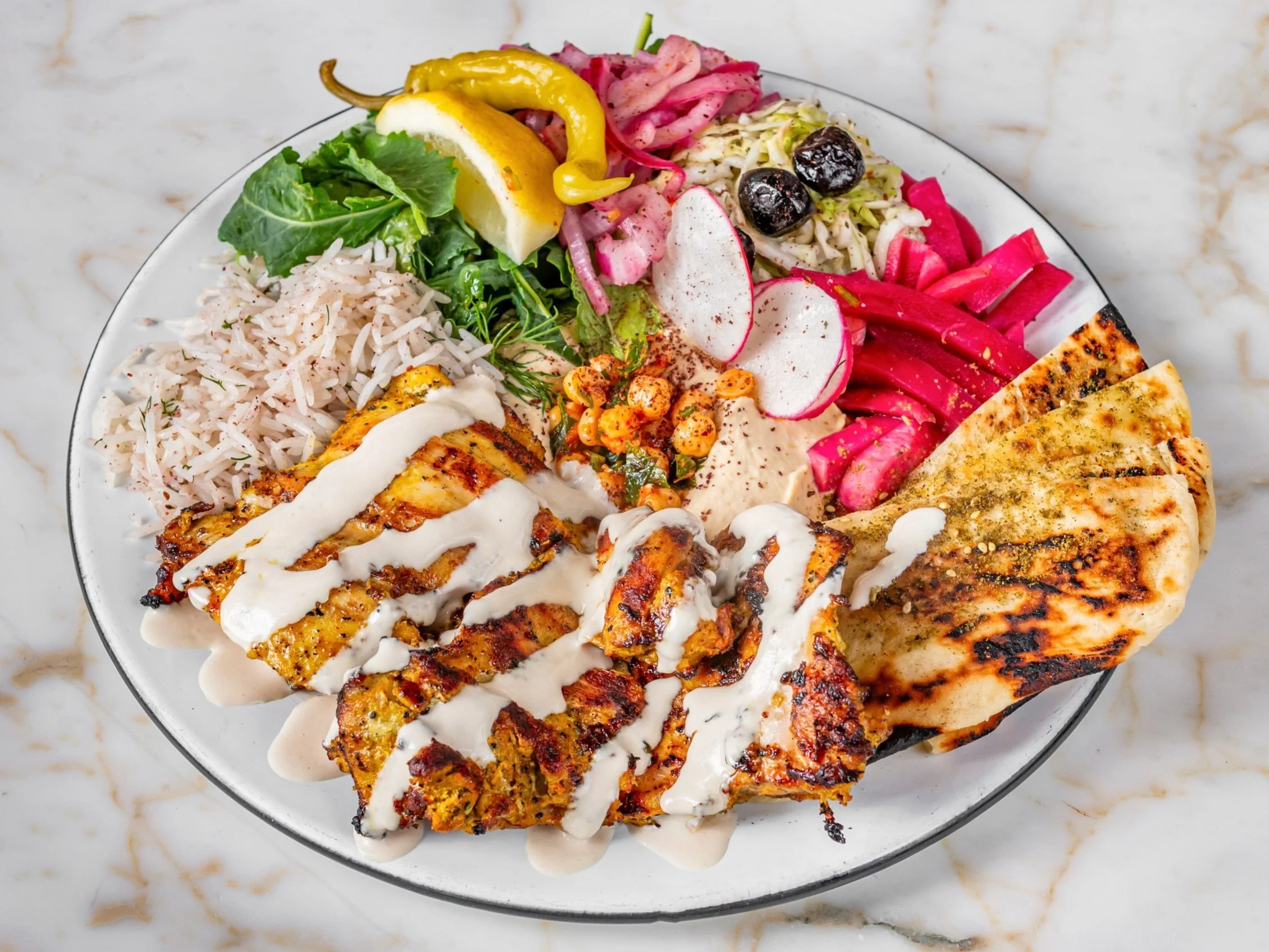 A colorful plate of various Mediterranean dishes including grilled chicken with white sauce, rice, pita bread, and assorted vegetables like pickles, radishes, and cabbage salads.