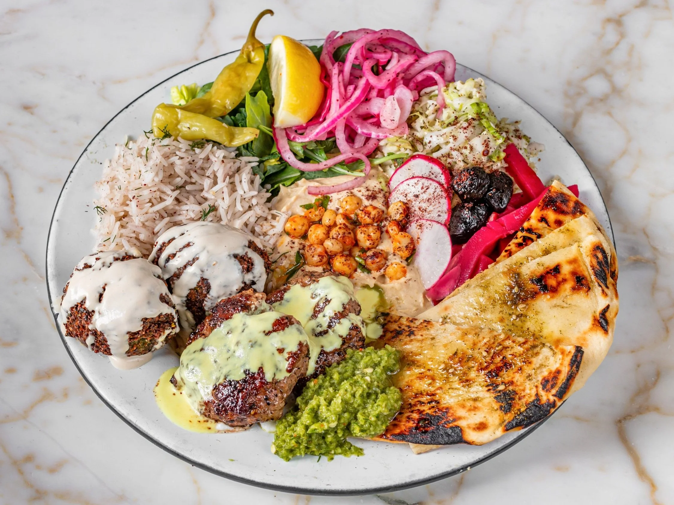 Plate of Middle Eastern dishes including rice, grilled meat with sauce, falafel, pita bread, hummus, tabbouleh, pickles, and assorted salads on a marble surface.