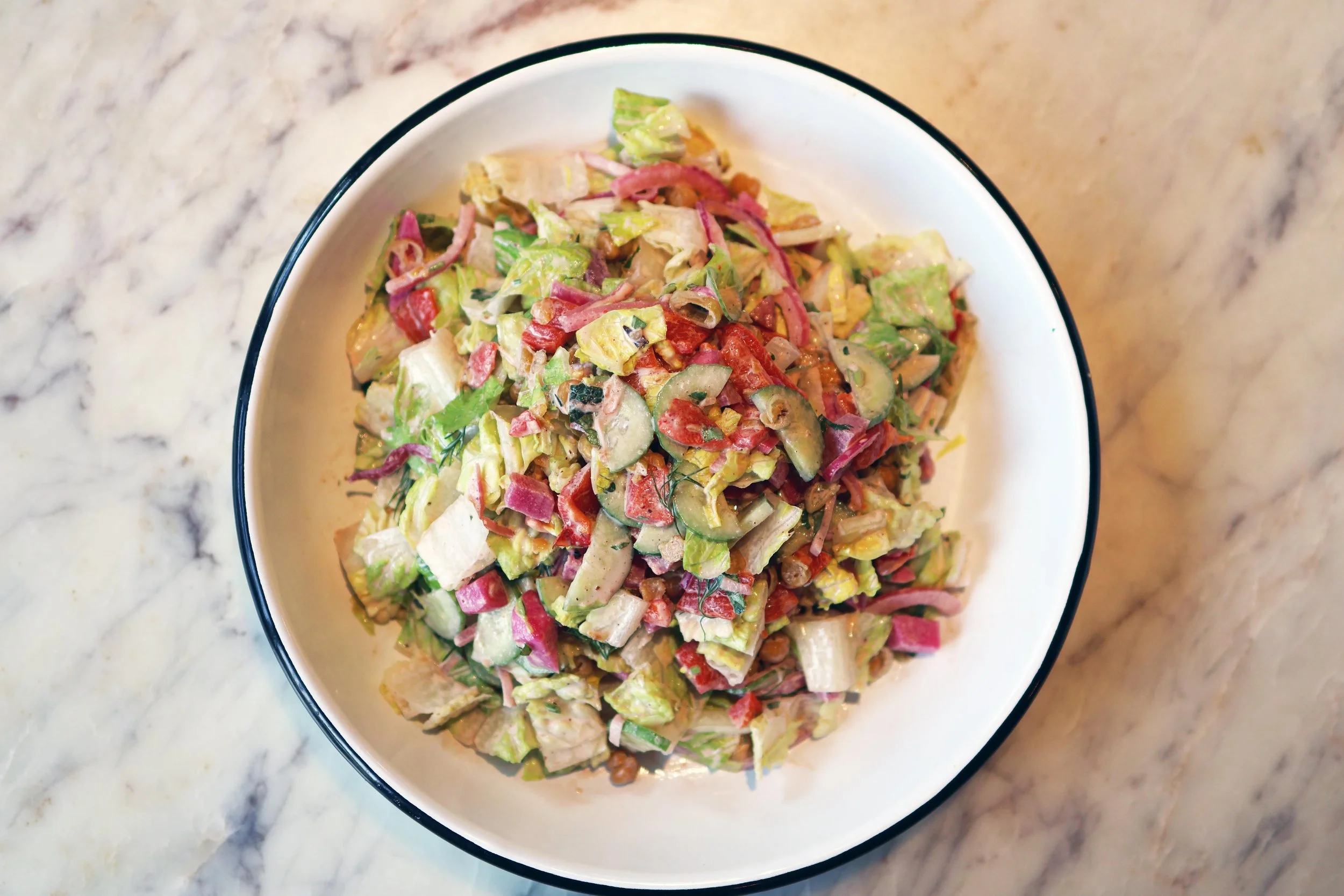 Bowl of chopped vegetable salad with lettuce, cucumbers, tomatoes, radishes, peppers, and herbs on a marble surface.