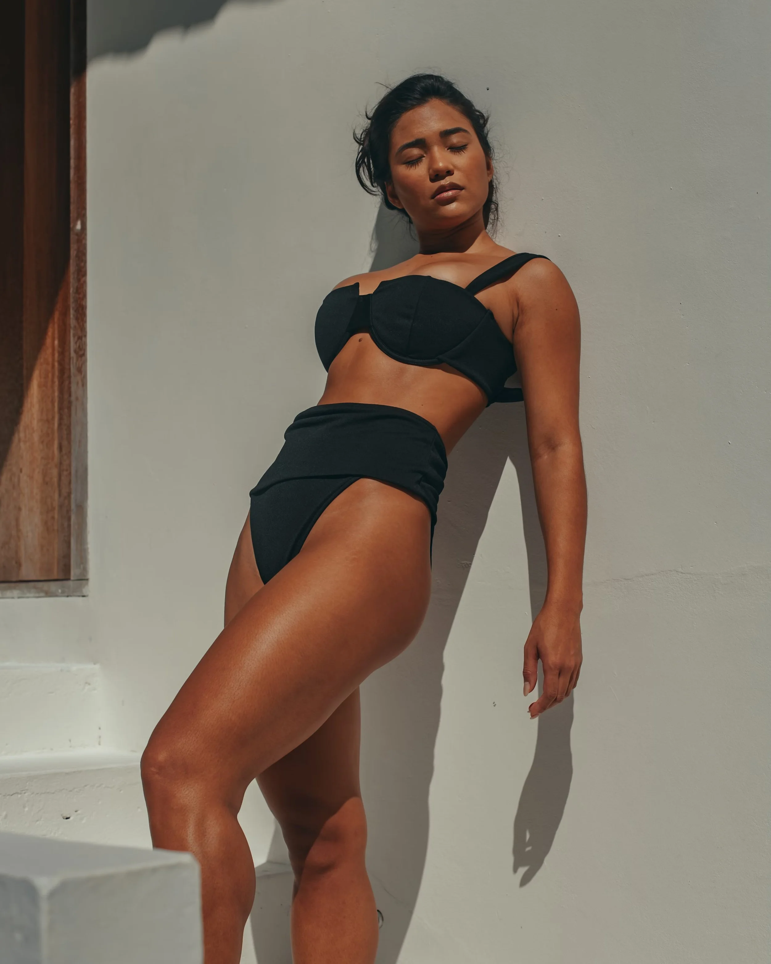 Person wearing a black bikini, standing against a white wall in sunlight, with eyes closed.