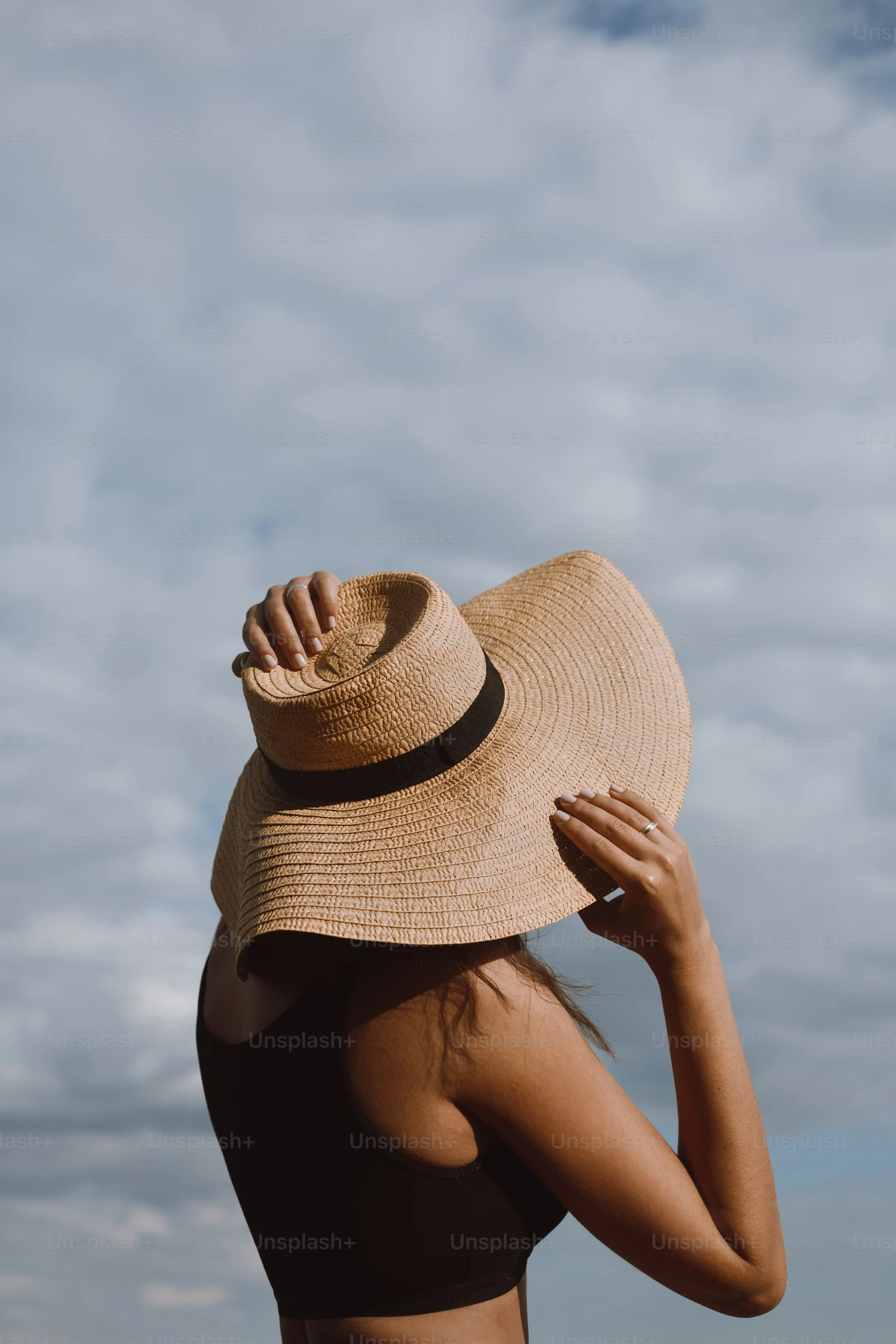 Woman in a black top wearing a large straw hat against a cloudy sky.