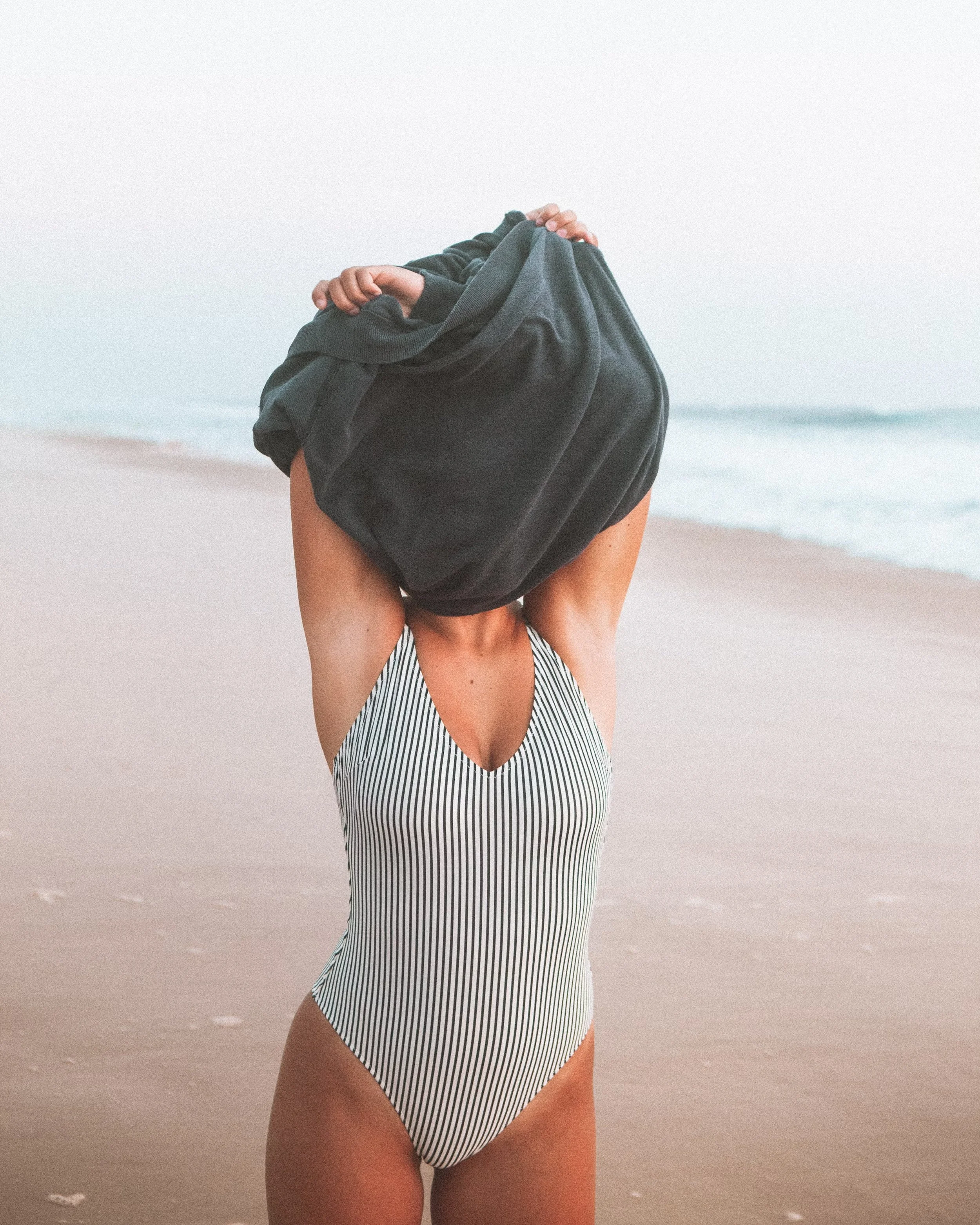 Person on a beach wearing a striped swimsuit, lifting a dark-colored shirt over their head.