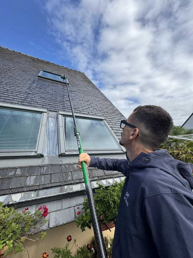 Utilisation d'une perche télescopique pour nettoyer une fenêtre située en hauteur sur la façade d'une maison à toit en ardoise. 