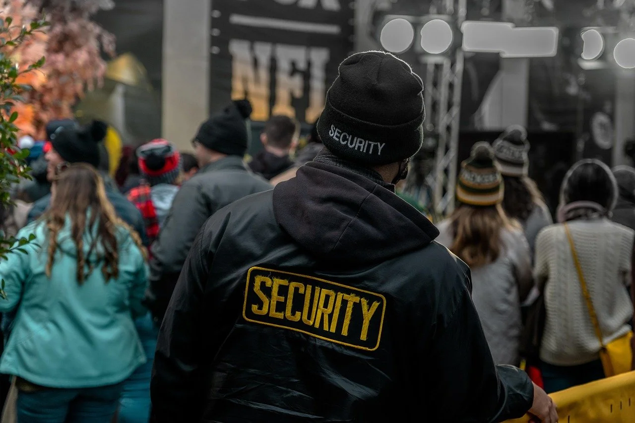 Security personnel monitoring a crowd at an outdoor event with lights and signage in the background.