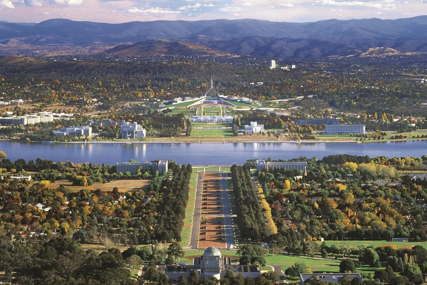 Aerial view of Canberra, Australia, showing the National War Memorial, tree-lined road, Lake Burley Griffin, and Parliament House, surrounded by buildings, greenery, and distant mountains.