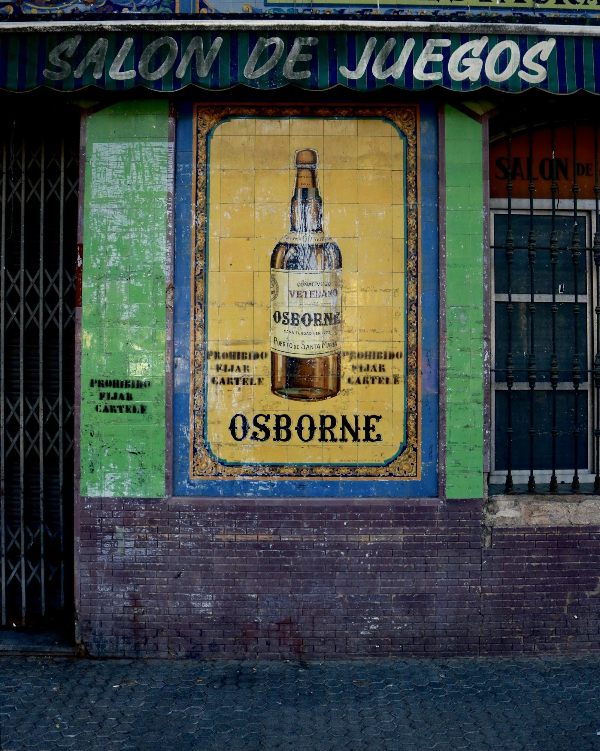 An outdoor sign for a game hall featuring a large, vintage-style advertisement of a bottle of Osborne brandy, with the text 'Salón de Juegos' at the top and 'Osborne' at the bottom, on a colorful tiled wall.