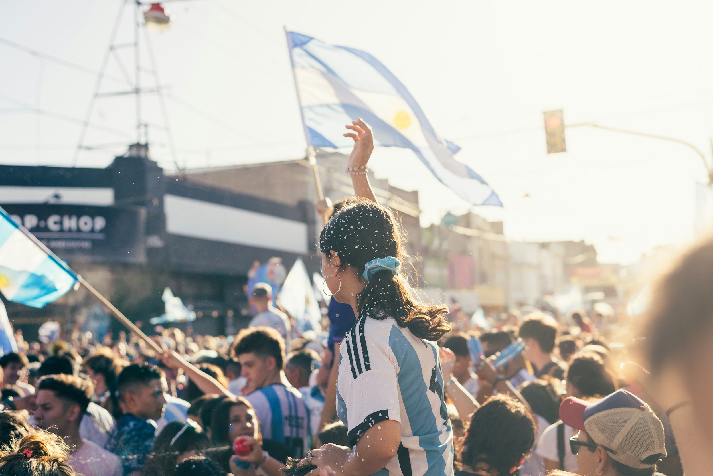 Crowd of people celebrating outdoors, with a woman in foreground wearing a sports jersey and a blue hair tie, waving a flag, flags visible in the background, sunlight streaming through.