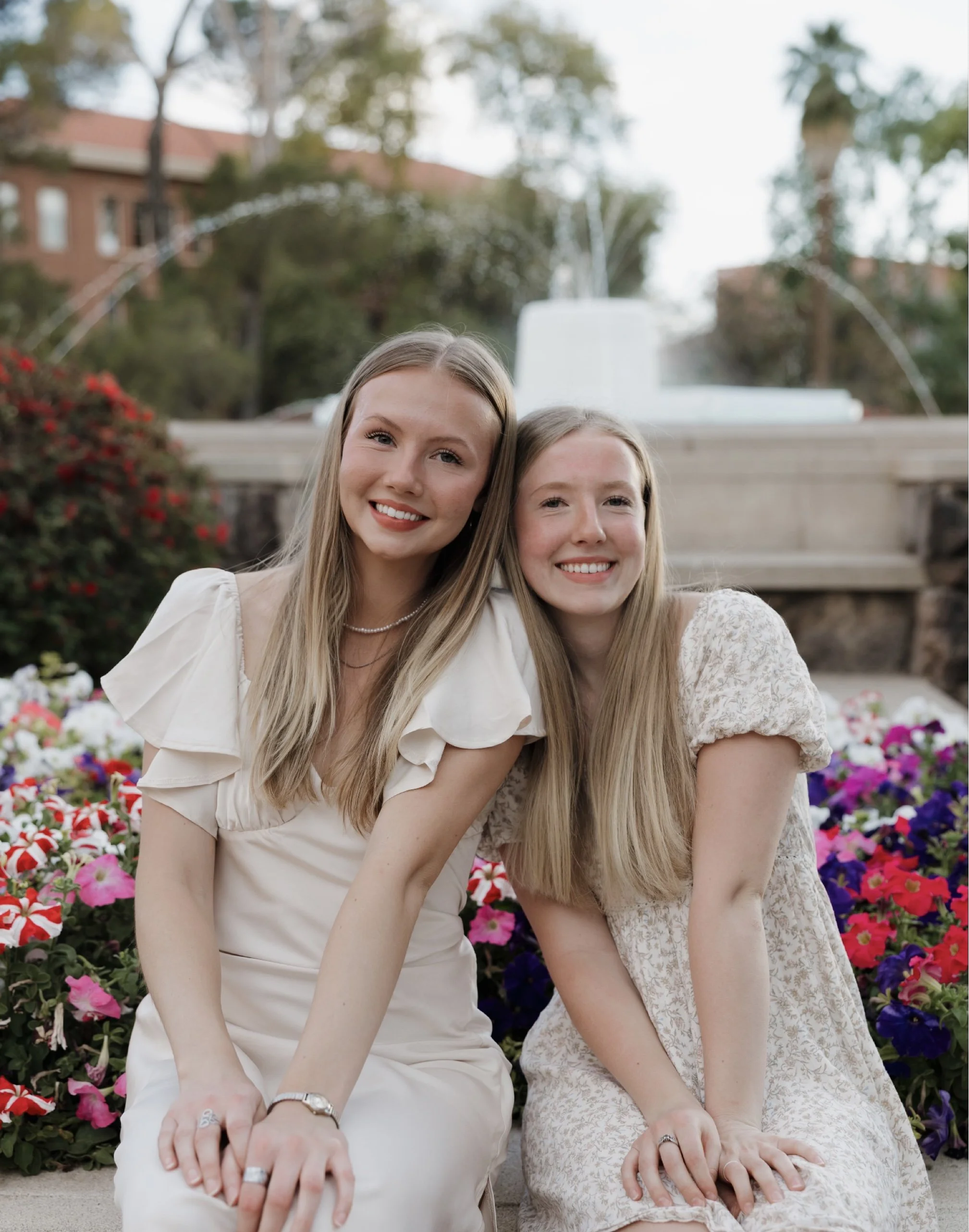 Two young women with long blonde hair sitting on the ground in front of a flower bed, smiling at the camera. Behind them, there is a fountain and trees.