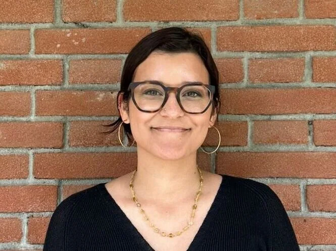 Portrait of a woman with short dark hair and glasses, wearing hoop earrings and a gold necklace, smiling in front of a red brick wall.