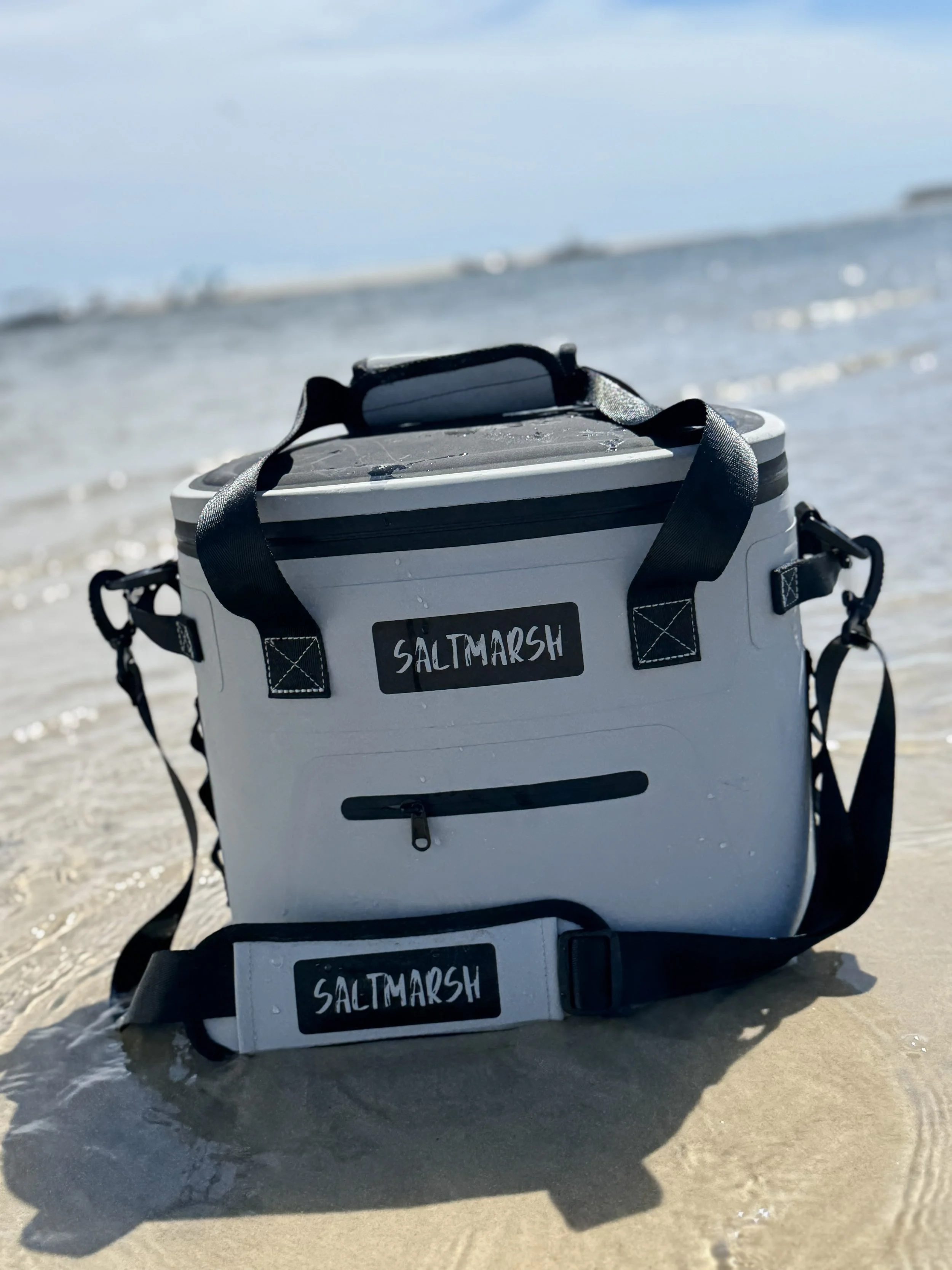 A white saltwater fishing cooler with black straps and a front zipper, sitting on wet sandy beach near the ocean, with the water and distant shoreline in the background.