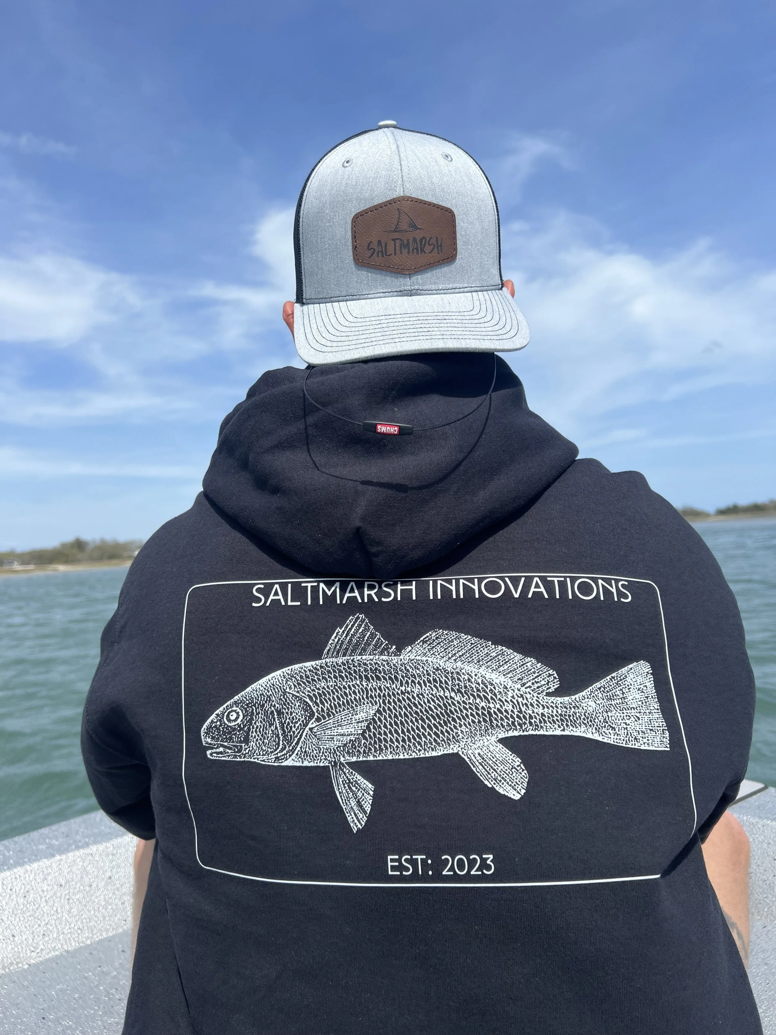 Person wearing Saltmarsh Innovations hoodie and baseball cap on a boat, with a water body and blue sky in the background.