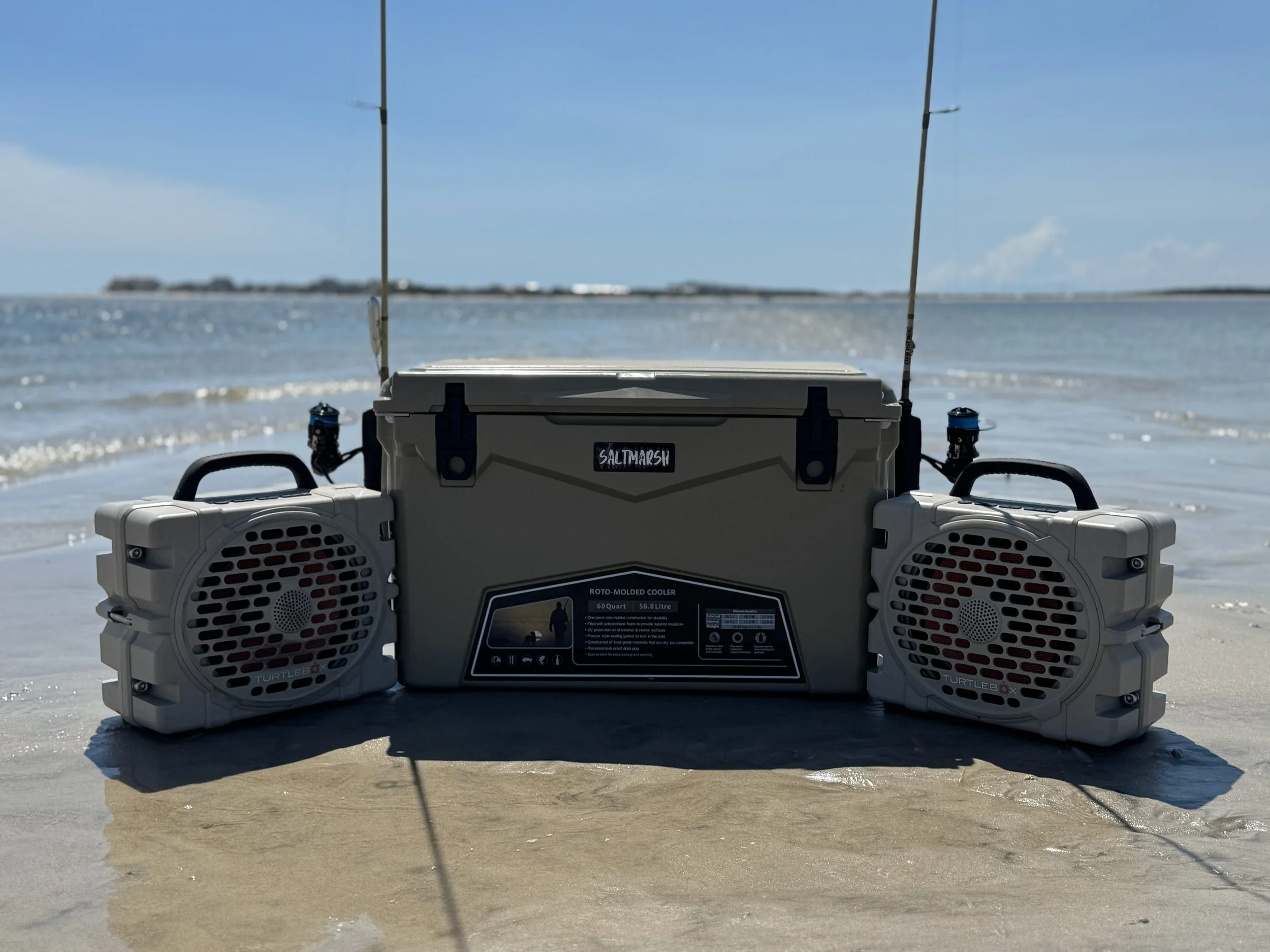 A portable saltmarsh cooler with two built-in speakers placed on the sand near water, with a calm ocean and a distant shoreline in the background under a clear blue sky.