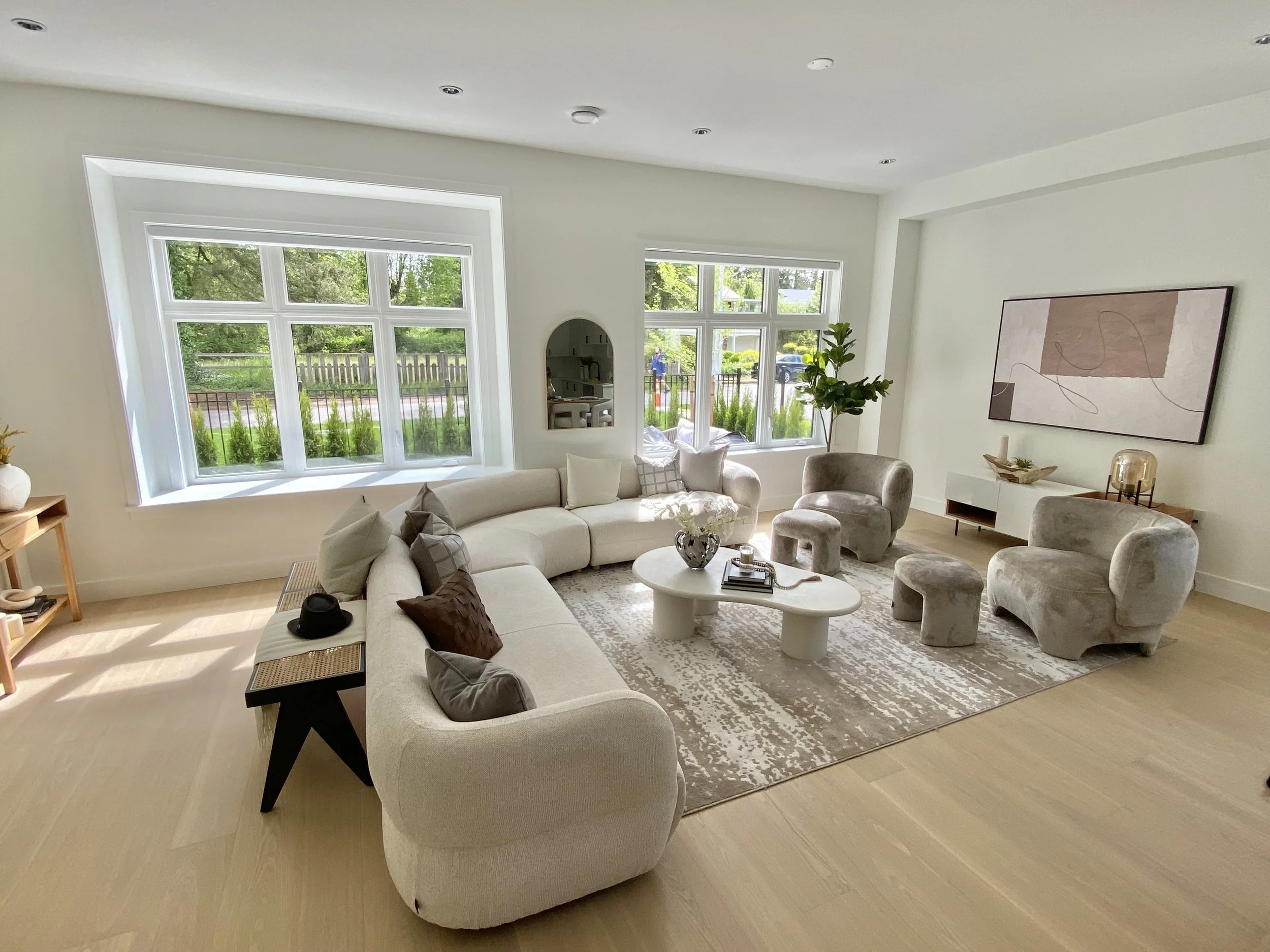 Bright living room with large windows, white L-shaped sofa, two beige armchairs, a white coffee table, and modern decor.