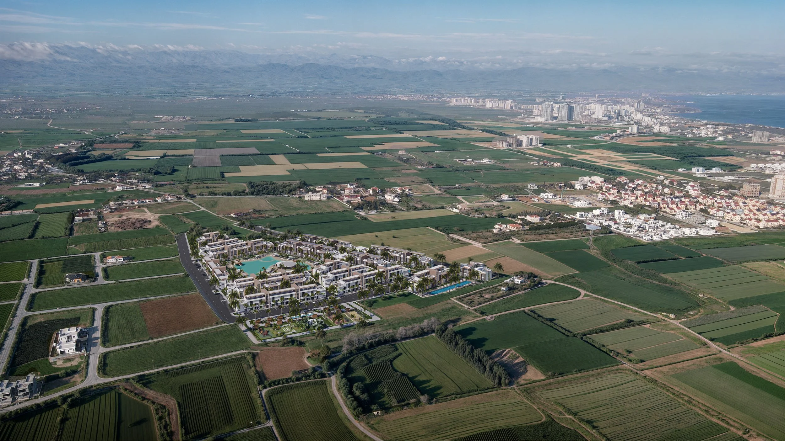 Aerial view of a residential complex with swimming pools surrounded by green fields and farmland, with mountains and a city skyline in the background.