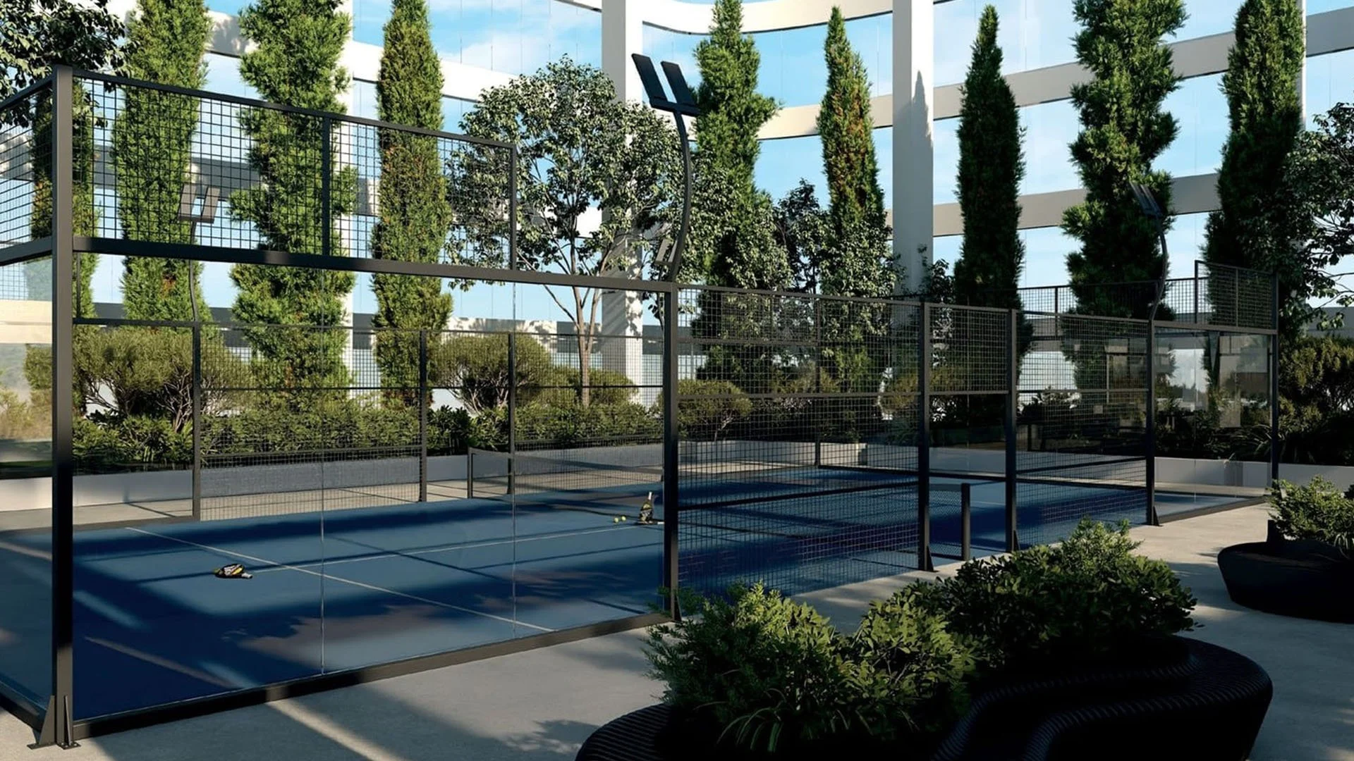 Empty outdoor padel tennis court with a black net and three paddles, surrounded by greenery and trees in an urban setting.