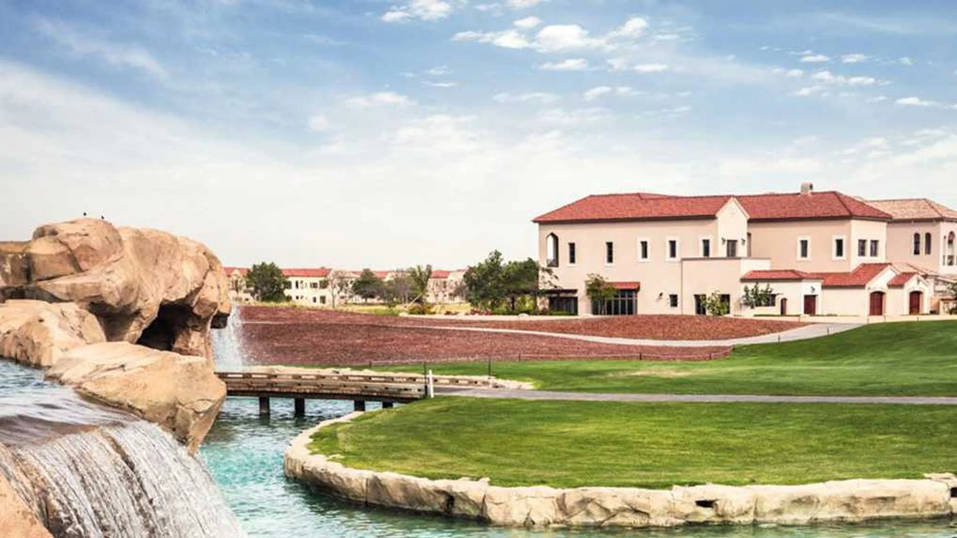 A landscaped area with a water feature, rocks, a small bridge, green grass, trees, and residential buildings with red-tiled roofs under a partly cloudy sky.
