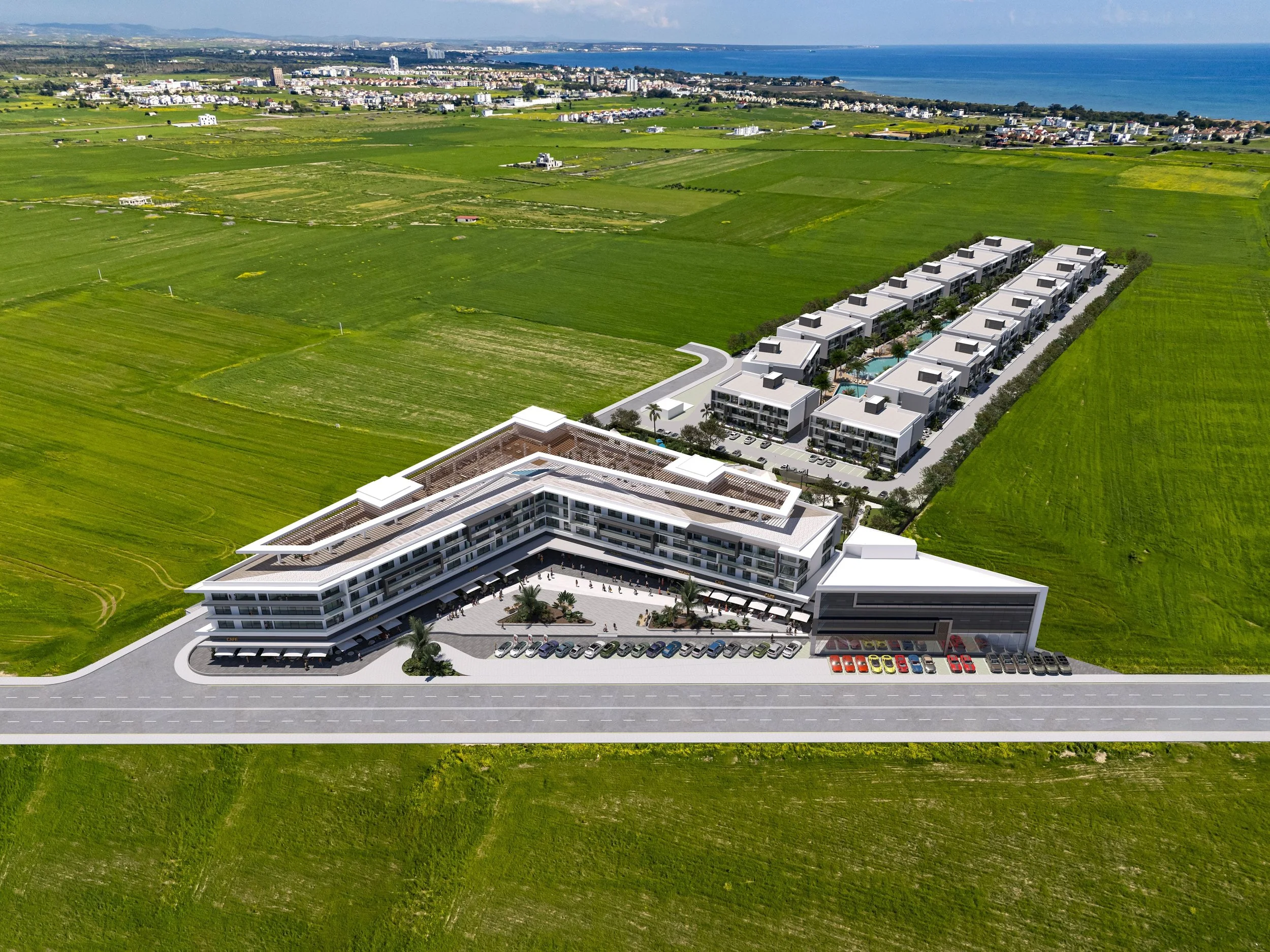 Aerial view of a modern hotel complex with multiple buildings, parking spaces, and a landscaped courtyard, surrounded by vast green fields and near the coast.