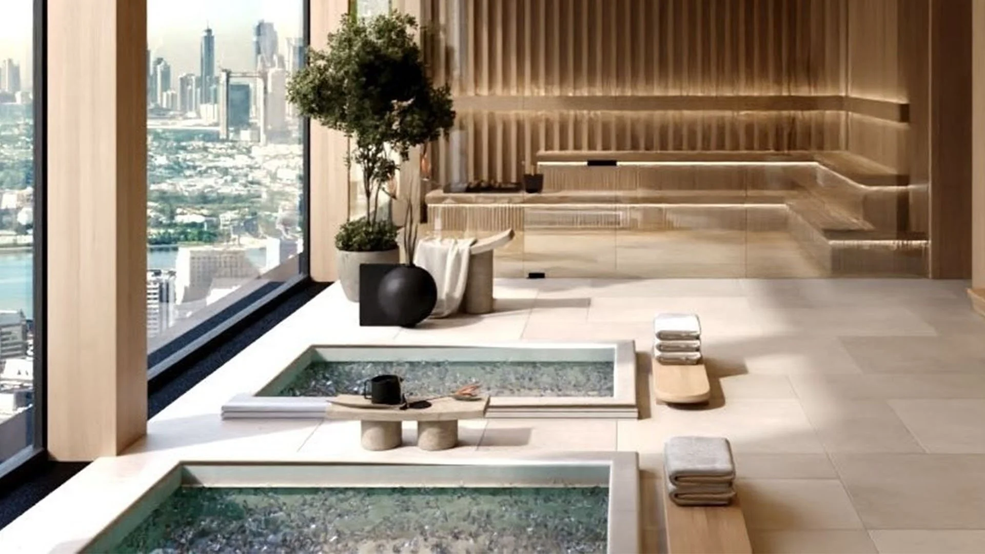 Modern indoor relaxation area with two small hot tubs, towels, a potted plant, and city skyline view through floor-to-ceiling windows.