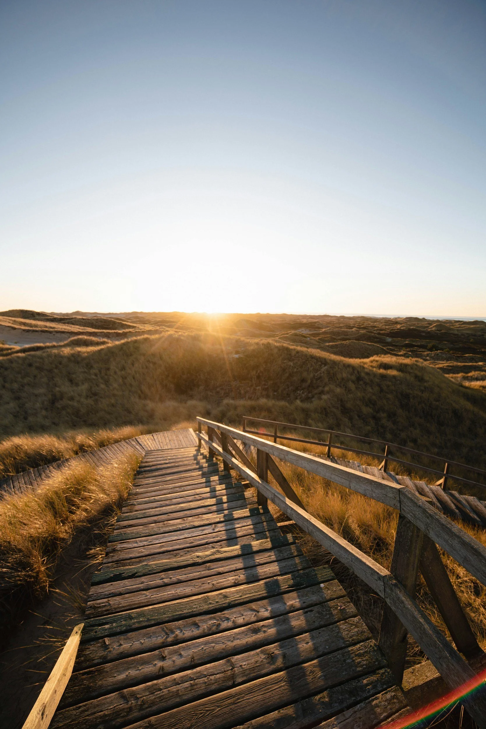 A wooden boardwalk leading over sand dunes at sunset, with the sun low on the horizon and casting long shadows.