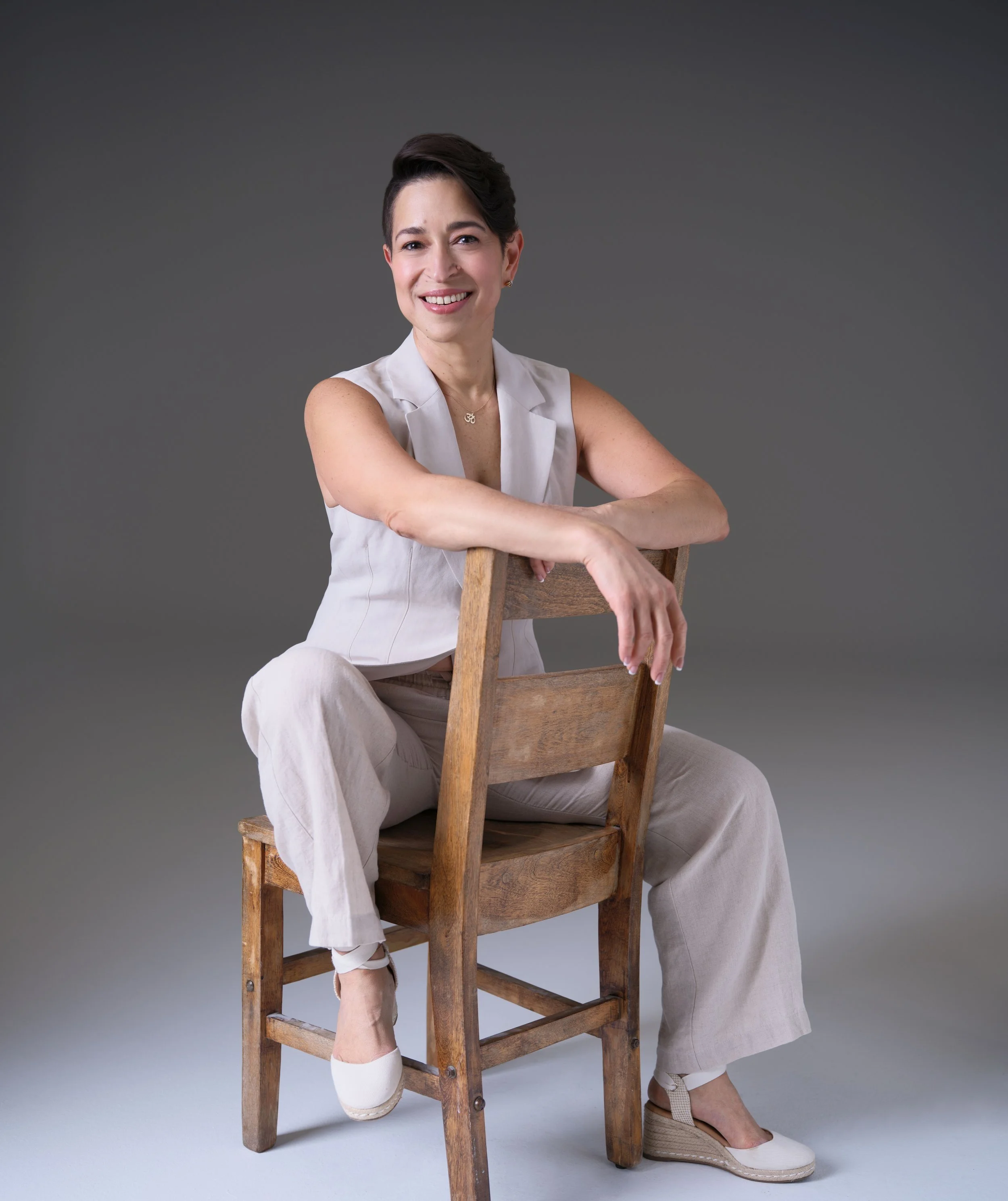 A woman with short dark hair, wearing a sleeveless light-colored top and light-colored pants, sitting on a wooden chair with one arm resting on the backrest and crossing her legs at the ankles, smiling against a plain gray background.