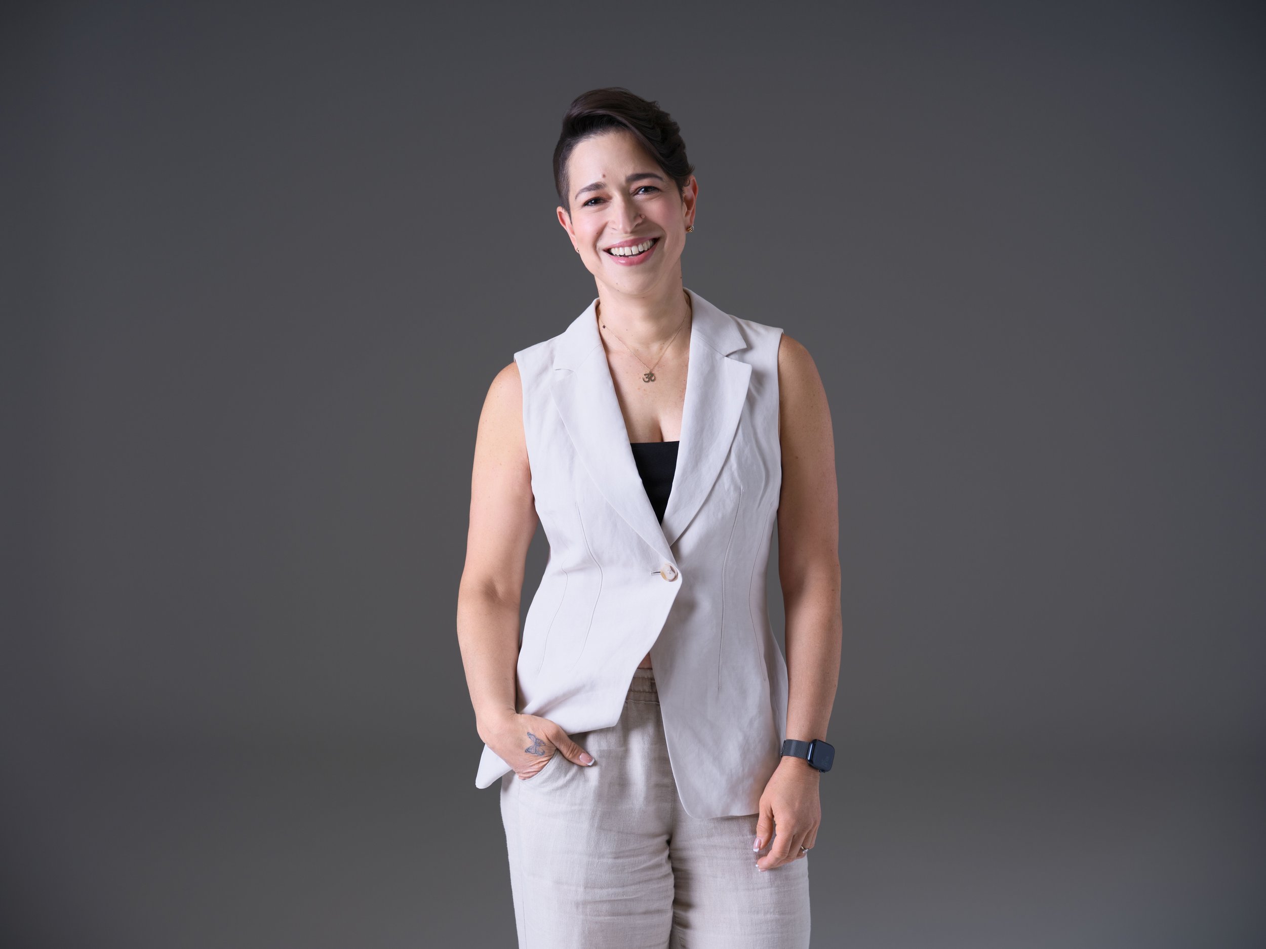 A woman with short dark hair, wearing a sleeveless white blazer over black top, smiling and standing with her hand in her pocket against a gray background.