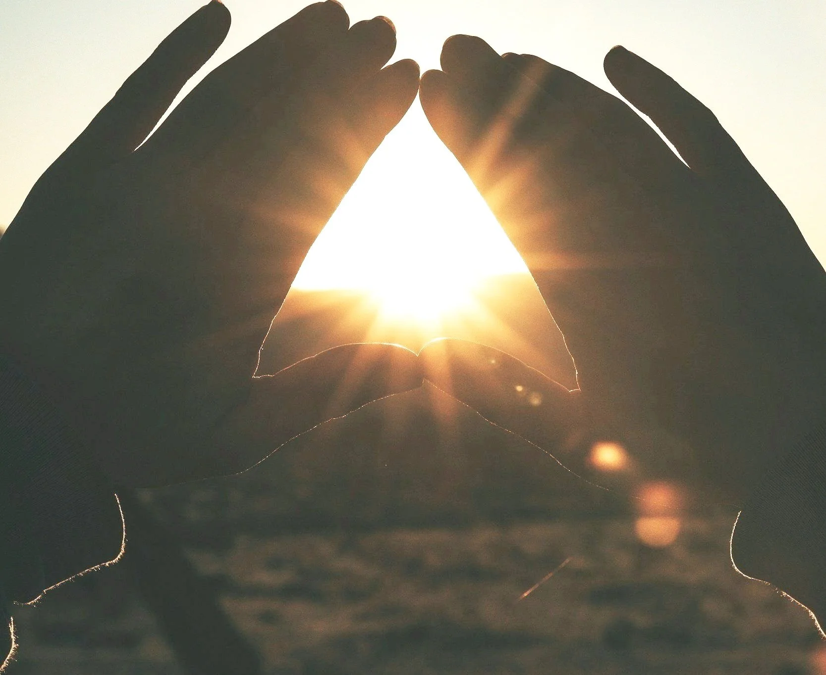 Hands forming a triangle shape framing the sun at sunrise or sunset.