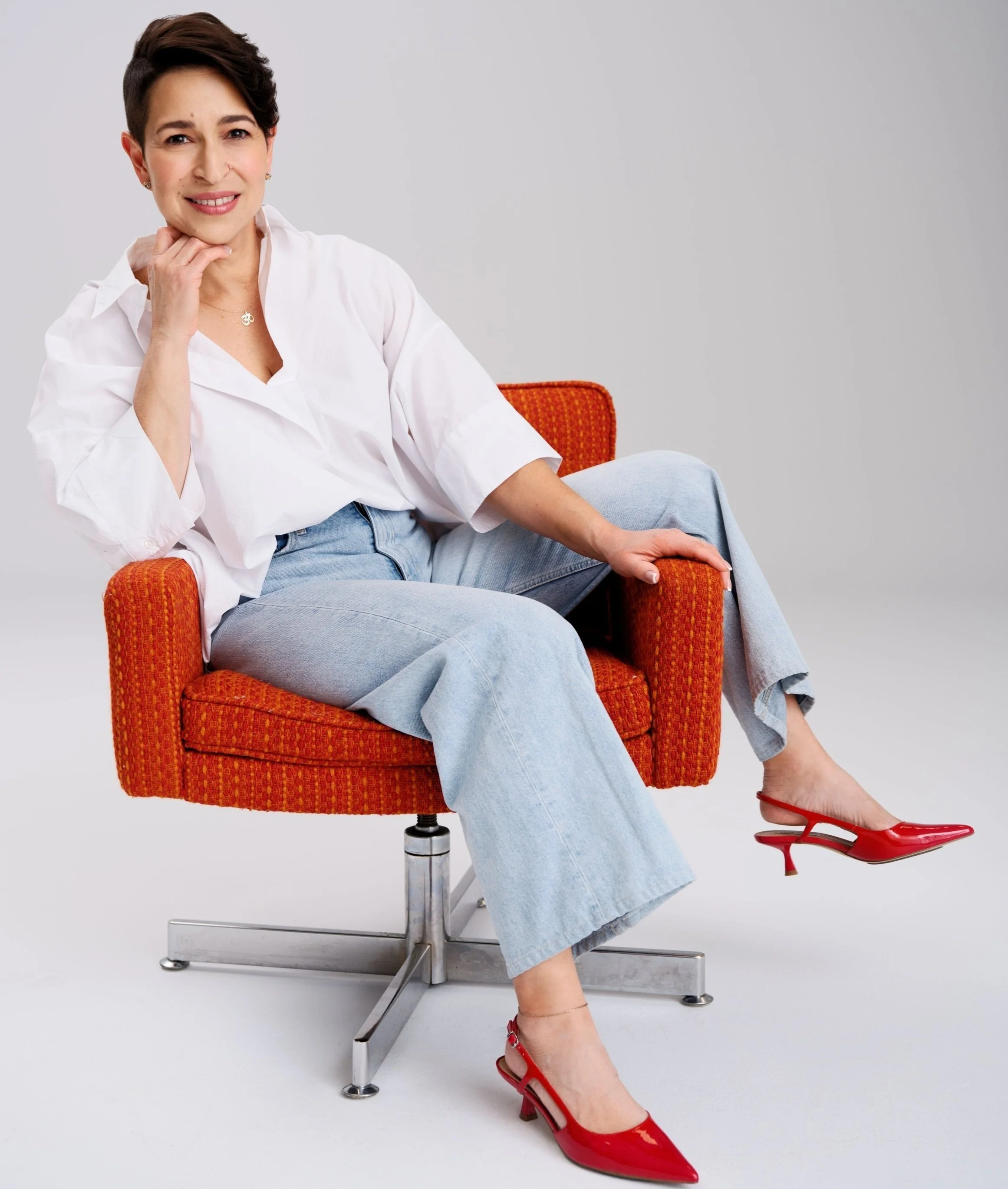 Woman sitting in an orange chair with one leg crossed over the other, wearing a white shirt, light blue jeans, and red high heels, against a plain background.