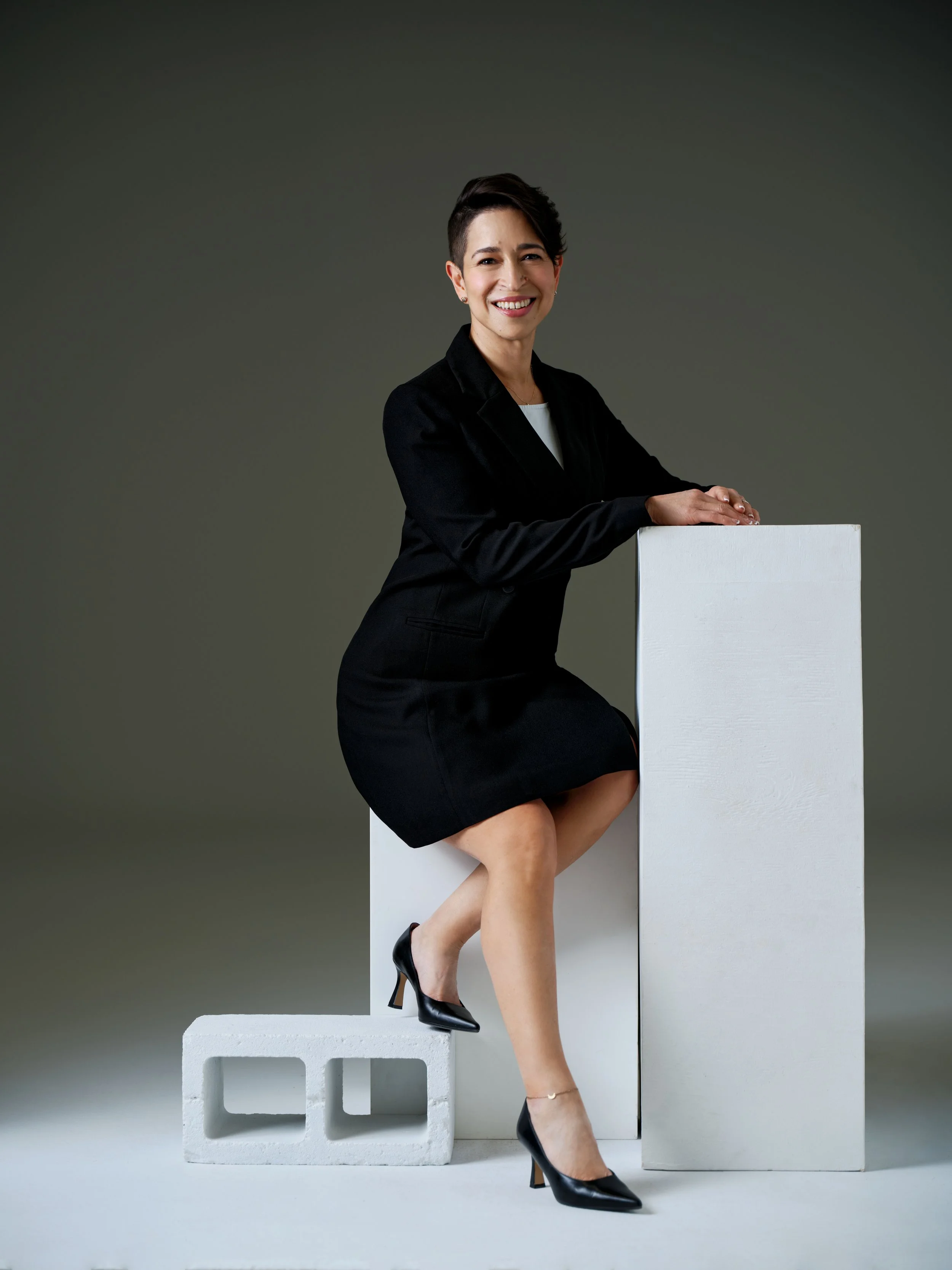 A woman dressed in a black business suit and high heels, smiling and posing with her hands on a white rectangular block, sitting with one foot on a gray cinder block.