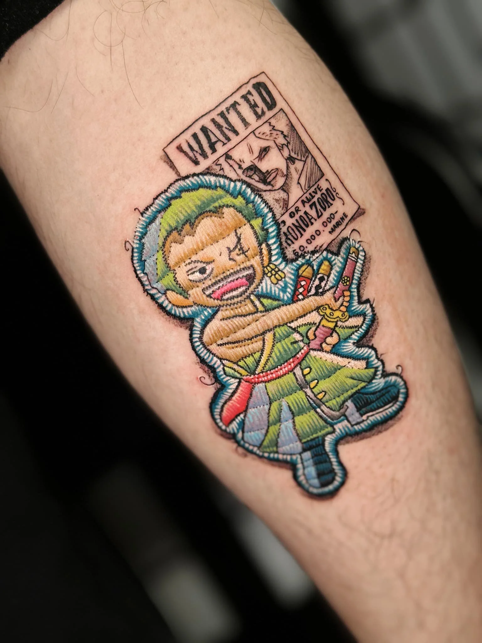 Colorful tattoo of a chibi-style character with green hair, a wink, and a finger pointing. Above is a manga-style panel with a fierce character and text reading 'WANTED' and a bounty notice. The tattoo features vibrant colors and bold black outlines.