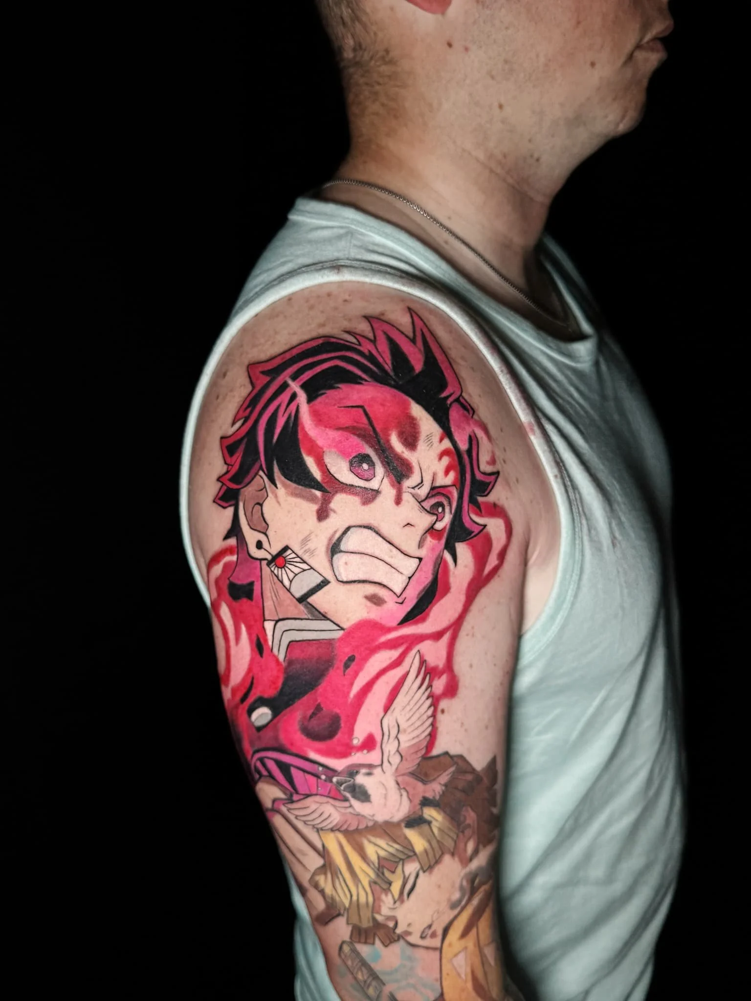 Colorful tattoo of a fierce anime-style character with black and pink hair, surrounded by red and pink flames, and a white bird on a branch on the person's upper arm.