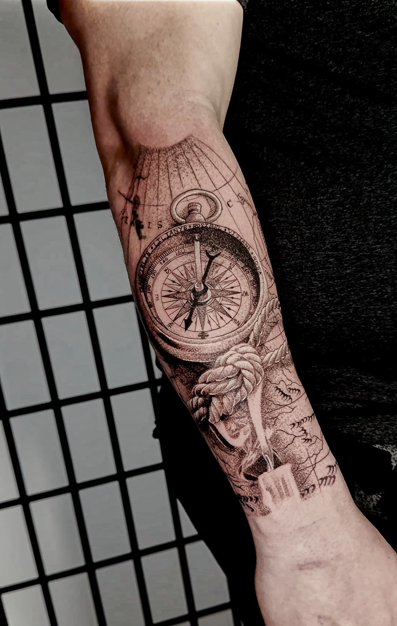Tattoo of a compass and a boat's anchor with nautical ropes on a person's forearm.